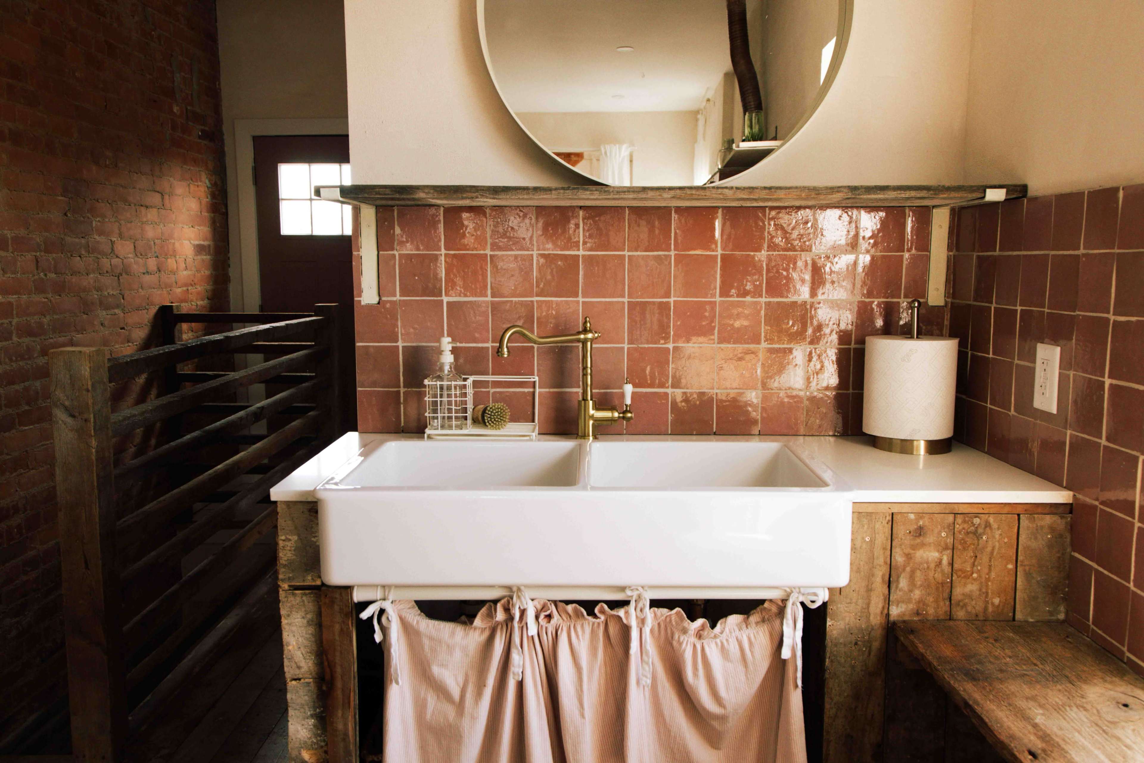 The image shows a stylish kitchen area featuring a white farmhouse sink with a gold faucet, surrounded by pink tiled walls and a wooden countertop, with a decorative lamp and curtain underneath the sink.