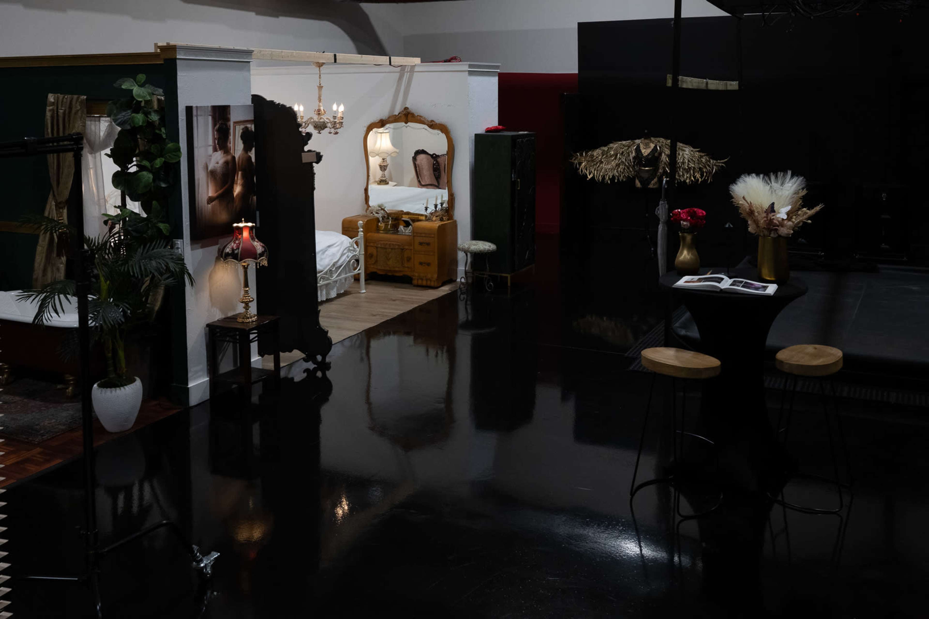 The image shows a studio set designed as a cozy interior space with a bed, decorative furniture, and plants, alongside a small table with flowers.