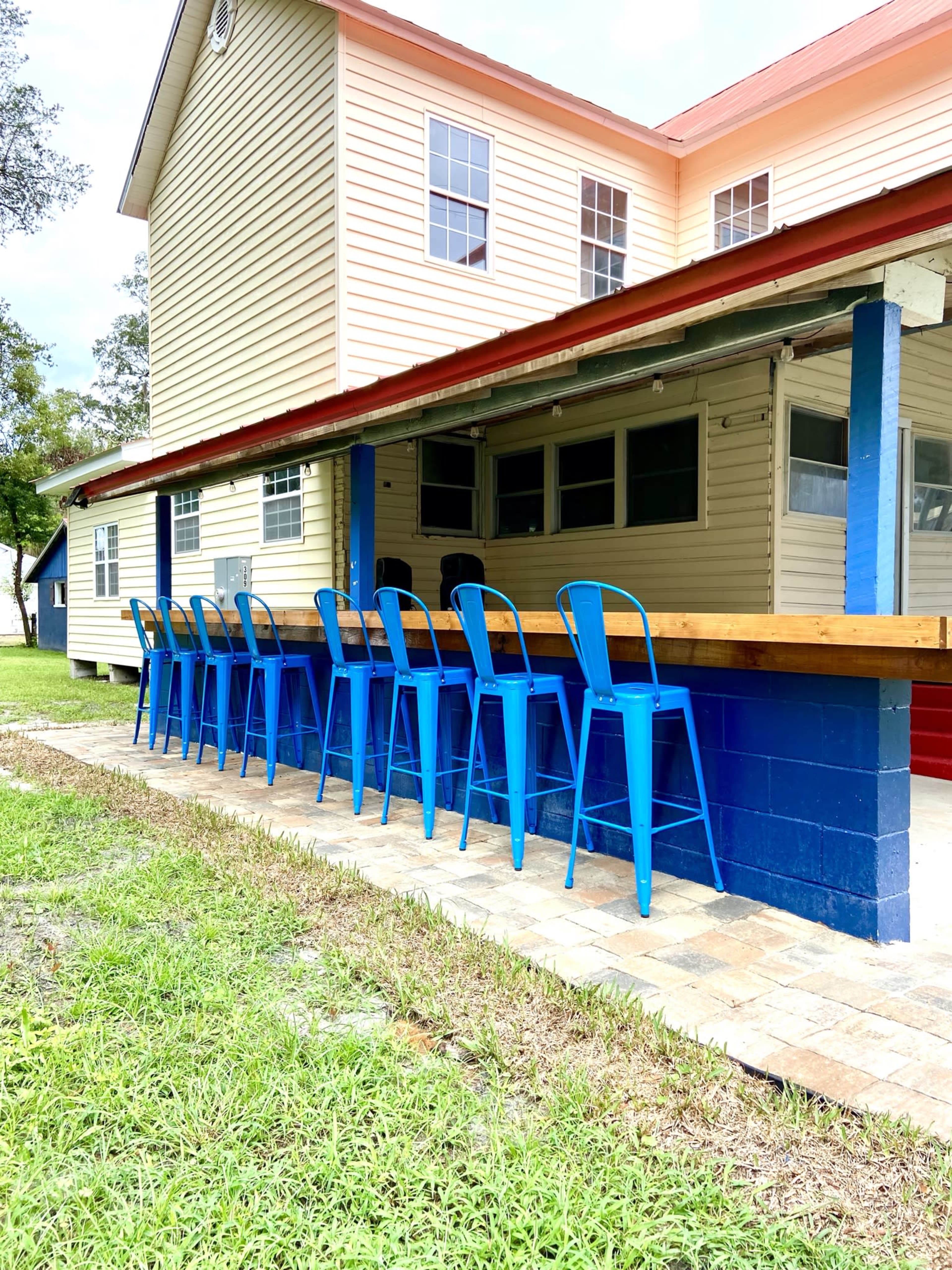 A row of blue metal bar stools is lined up in front of a wooden counter adjacent to a two-story yellow and blue building.