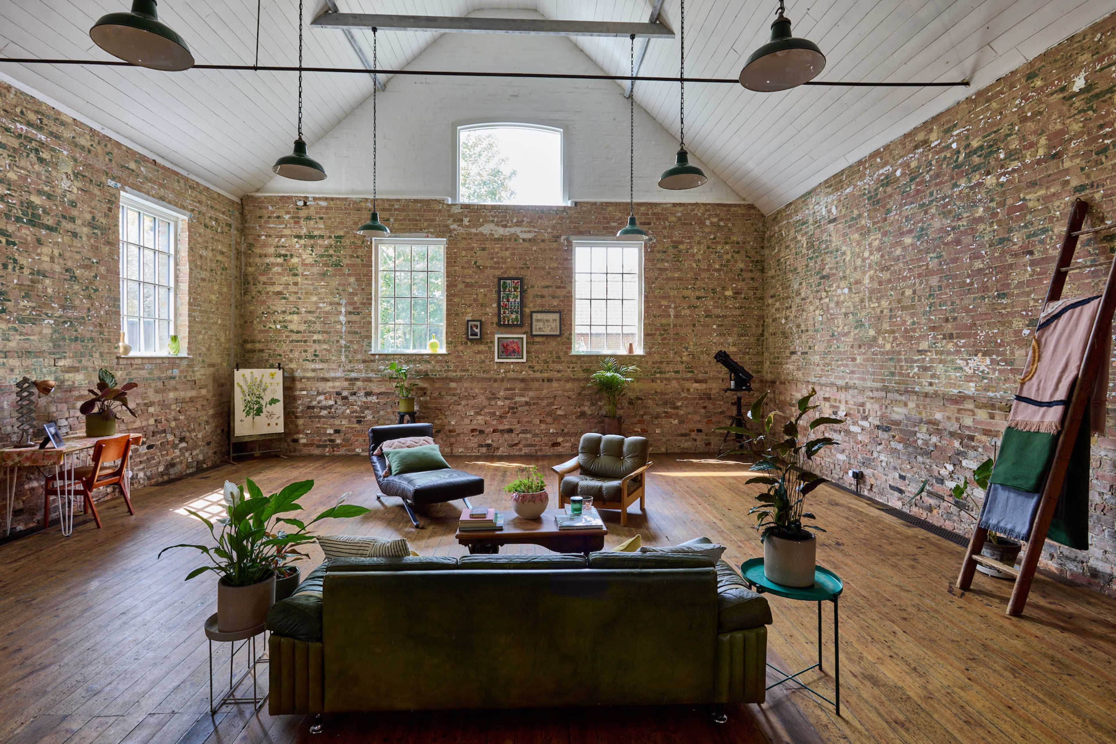 A spacious, brick-walled living area with large windows, wooden flooring, plants, and modern seating arrangements.