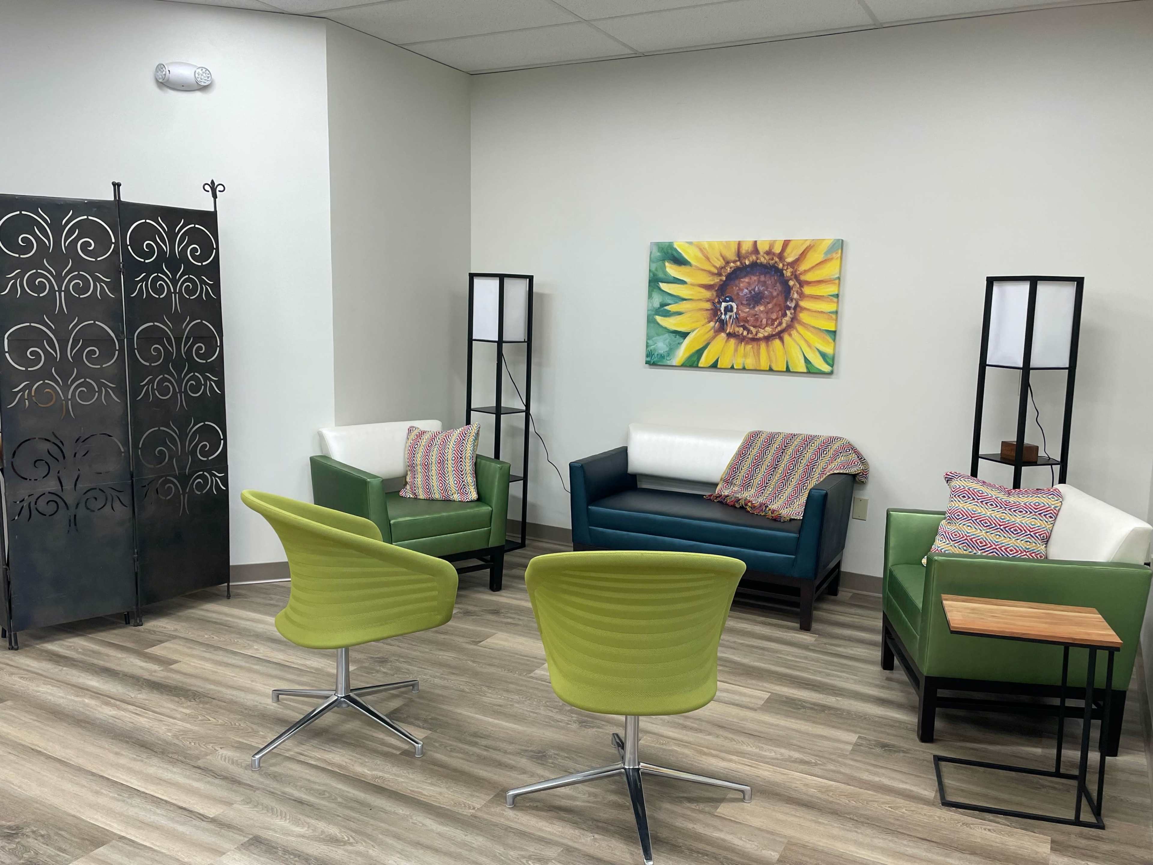 The image shows a waiting area with two green chairs and two couches, a decorative screen, and a sunflower painting on the wall.