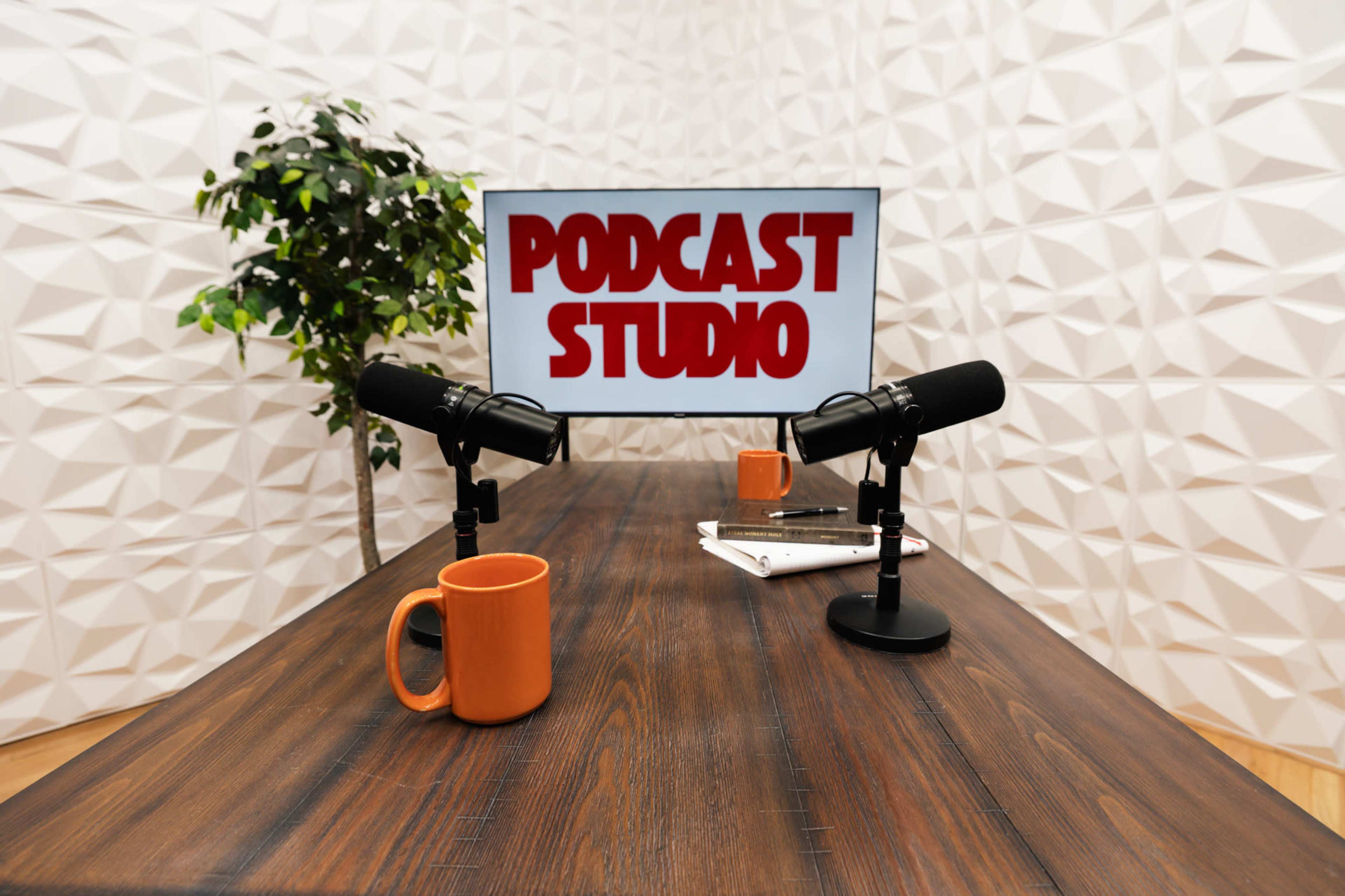 A minimalist podcast studio features a wooden table with two microphones, an orange mug, and a TV screen displaying the words "PODCAST STUDIO."