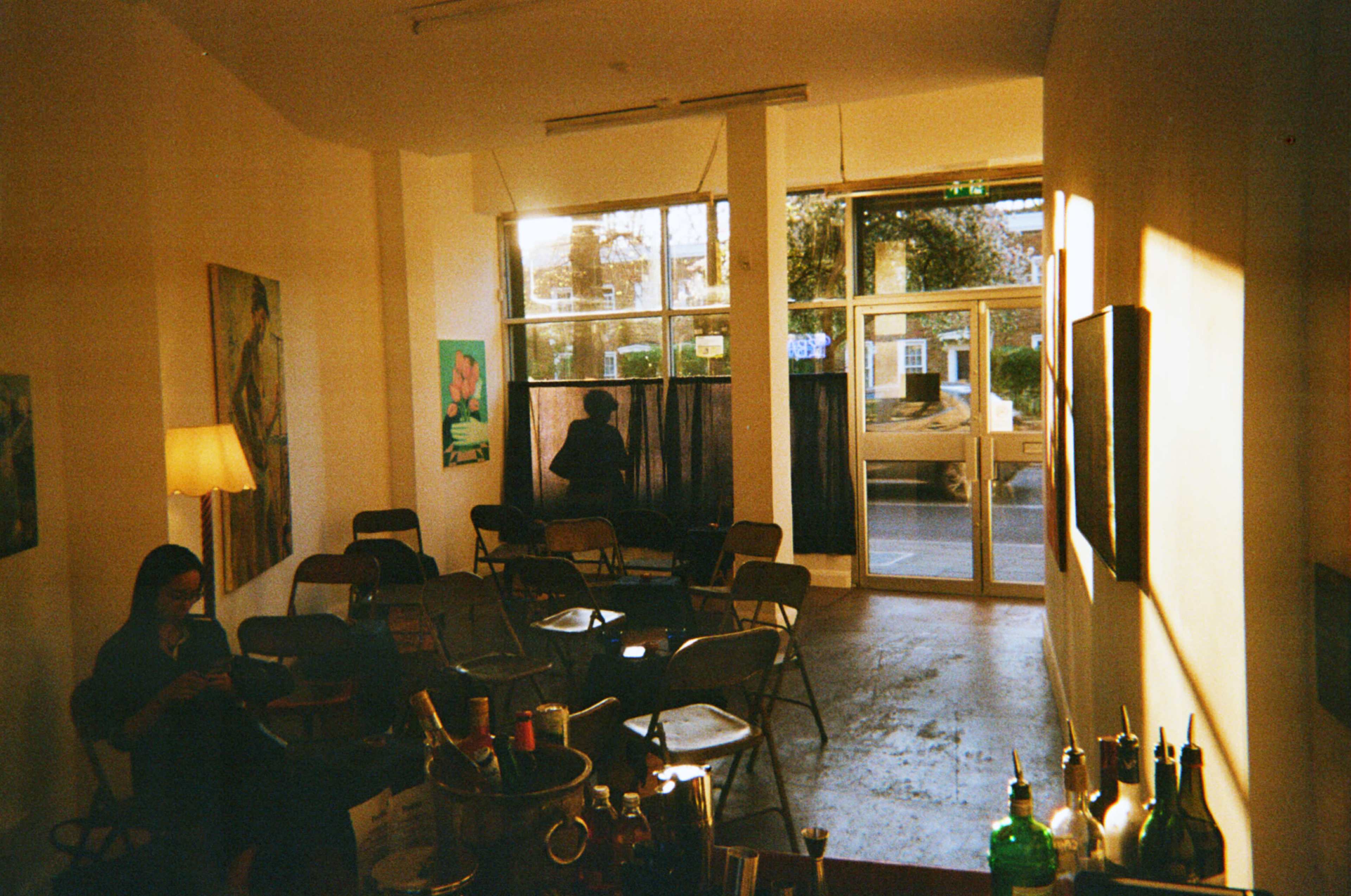 The image shows a sparsely furnished room with empty chairs arranged in rows, a floor lamp, and a person standing by the window.