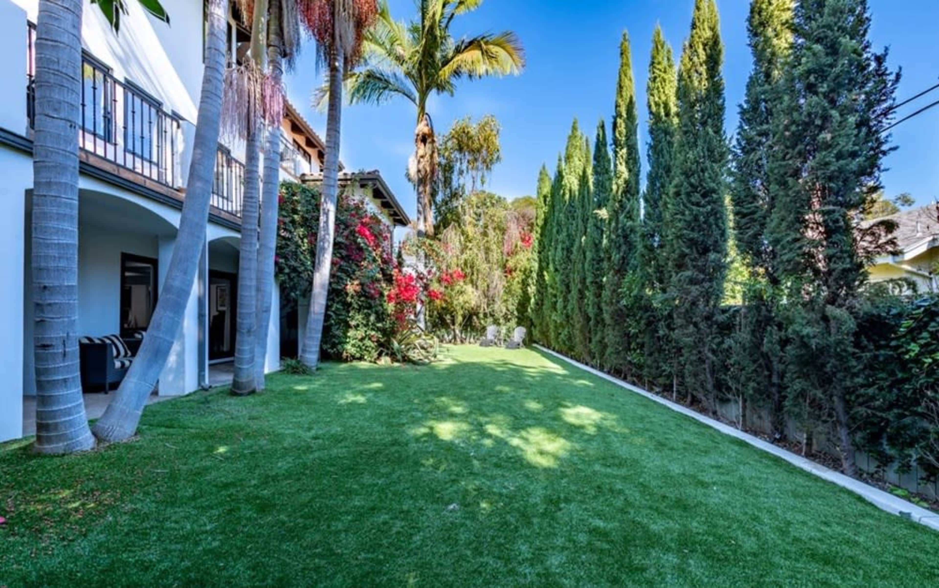 The image shows a well-maintained backyard with lush green grass, tall privacy trees, and vibrant flowering plants beside a white house.