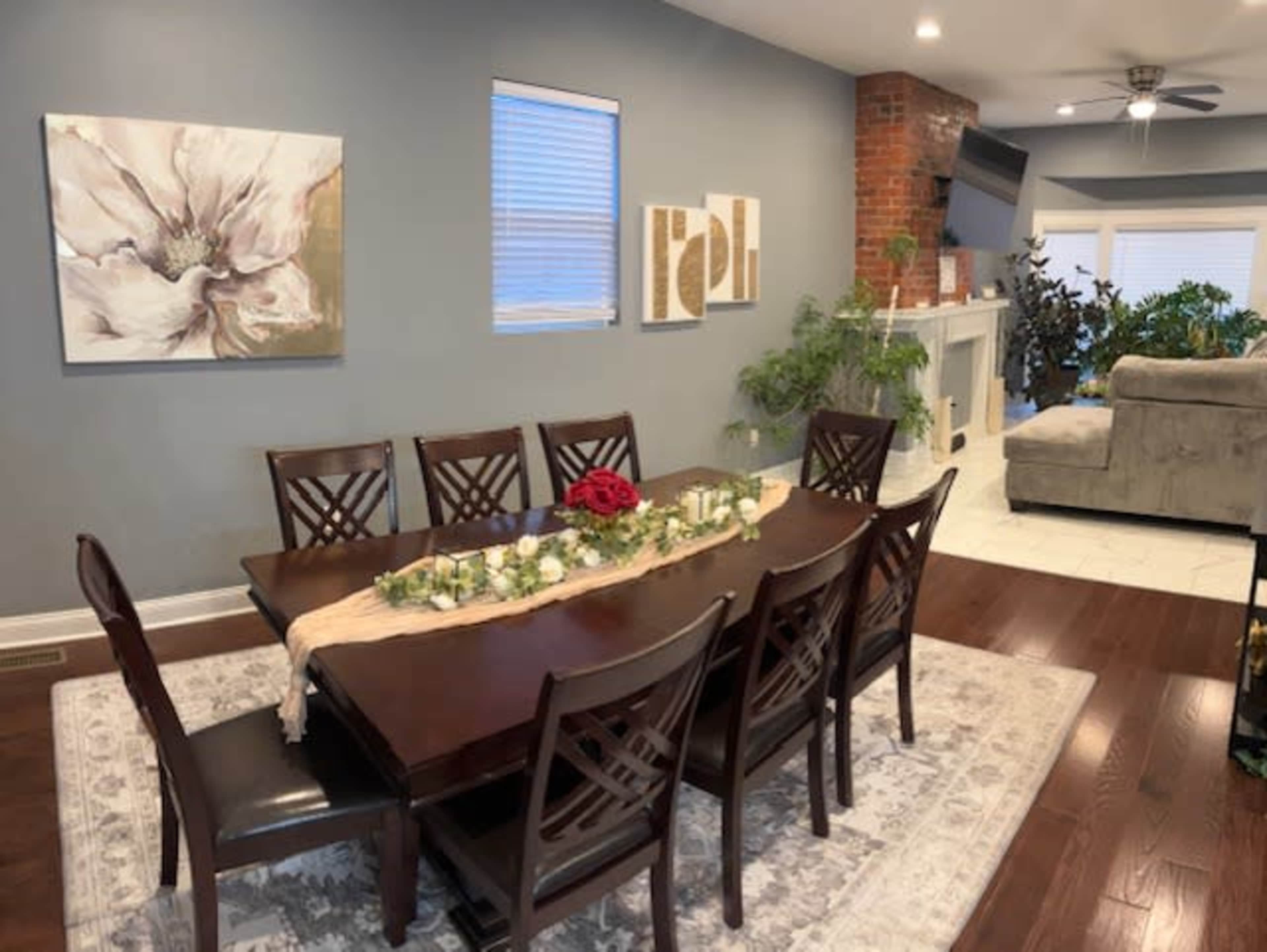 The image shows a dining area with a wooden table set for eight, surrounded by chairs, and featuring a decorative centerpiece, along with a seating area and plants in the background.