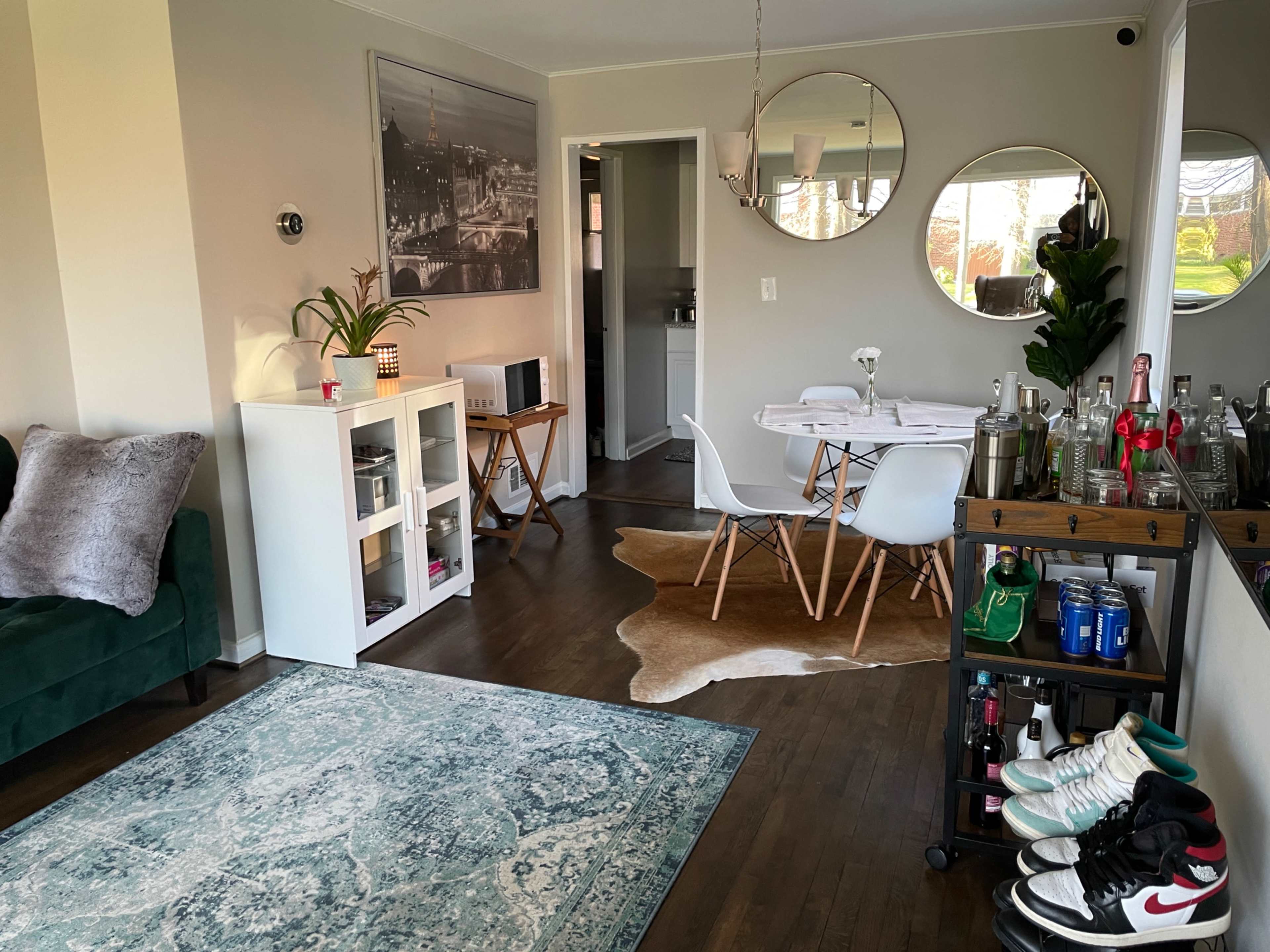 A modern living area features a green couch, a round dining table with white chairs, a bar cart, and decorative elements on the walls and floor.