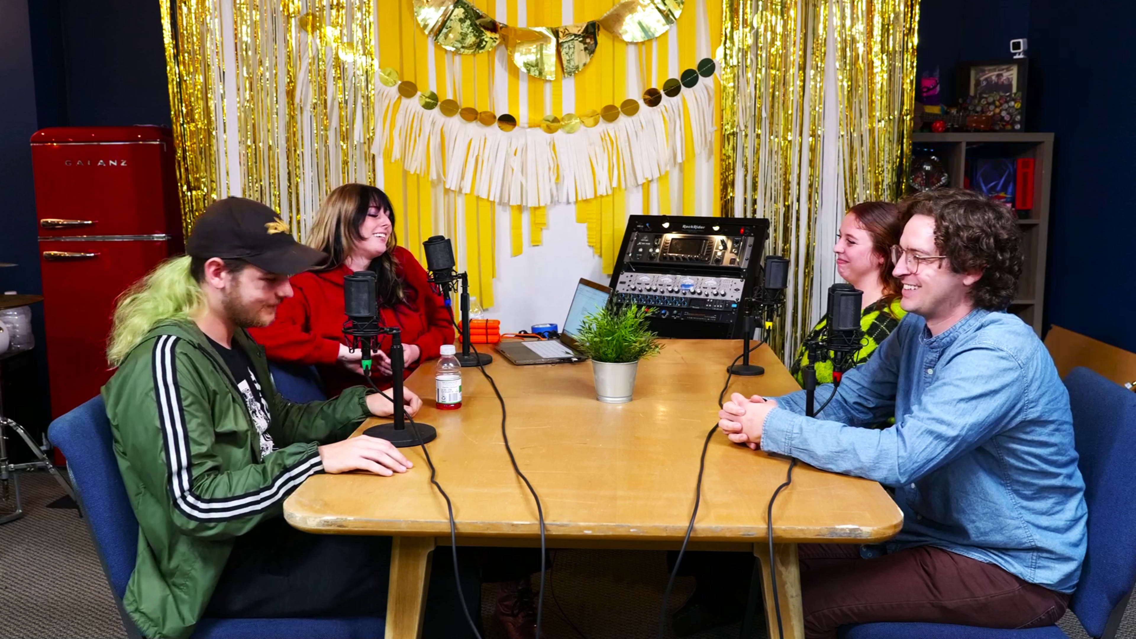A group of four people sits around a wooden table with microphones in front of them, surrounded by a brightly decorated backdrop of yellow stripes and metallic streamers.