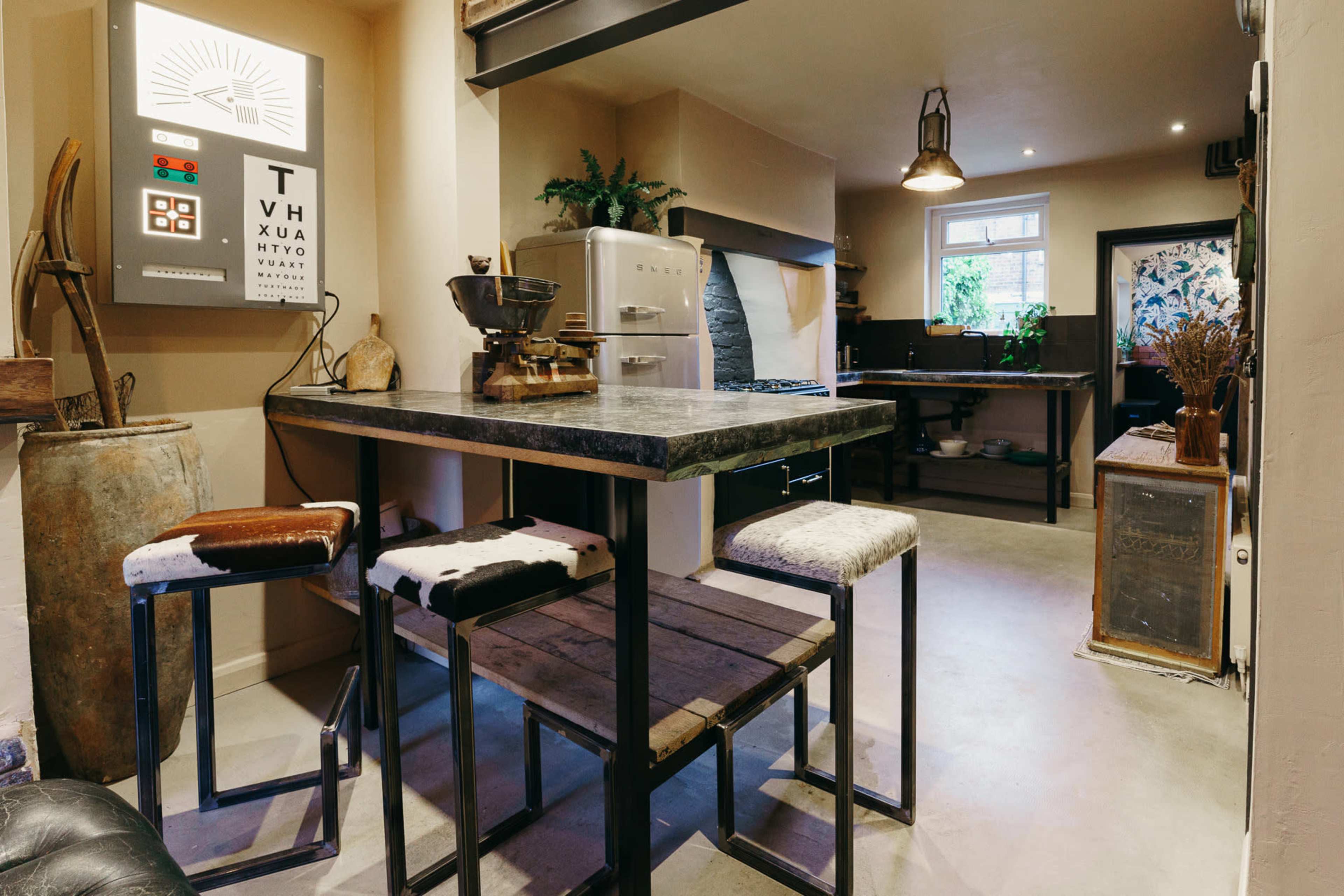 A rustic kitchen features a wooden table with four stools, a vintage stove, and a wall-mounted control panel.
