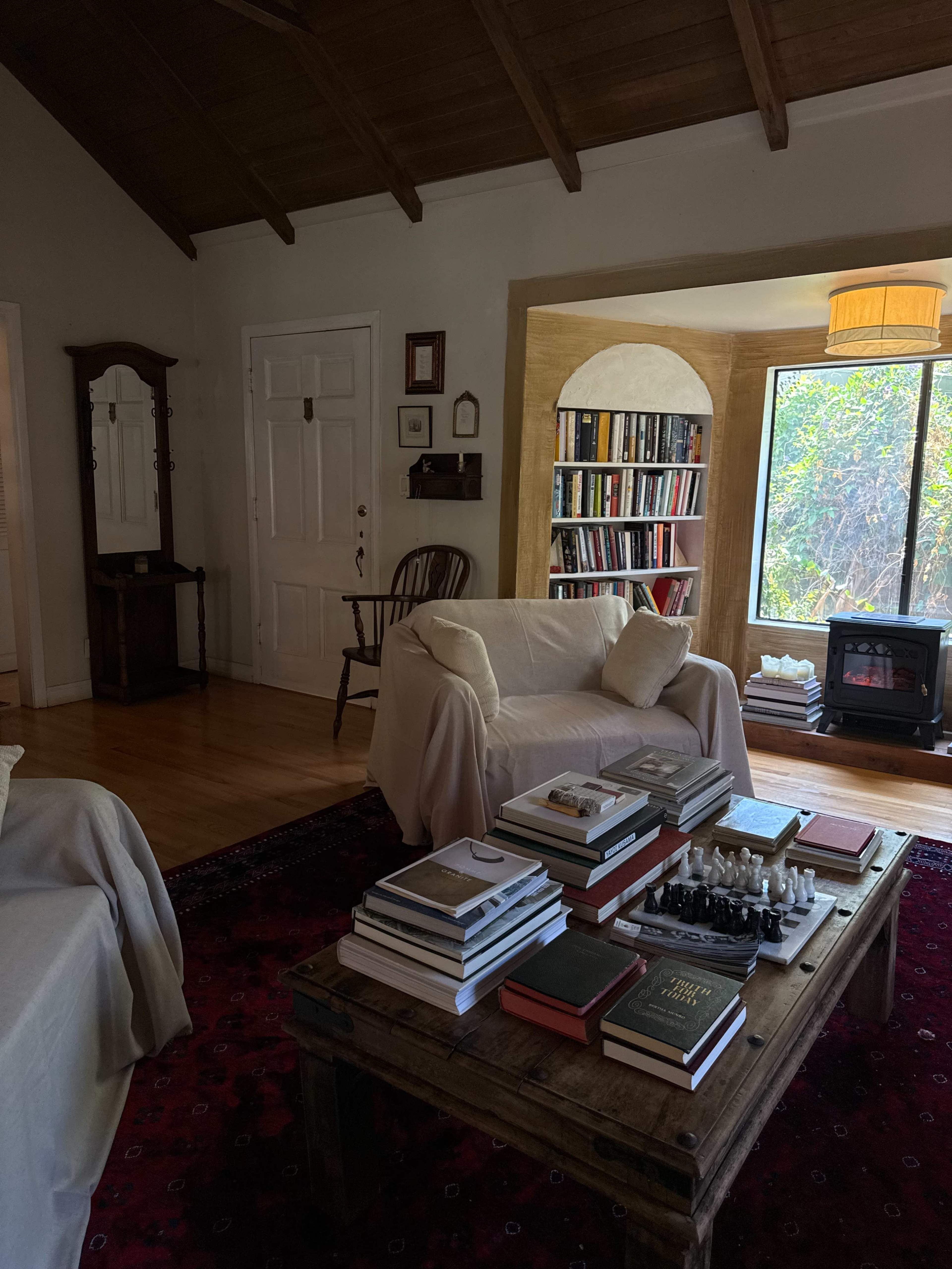 A living room features a coffee table stacked with books and a chess set, surrounded by a cozy seating area, a bookshelf filled with books, and a window letting in natural light.