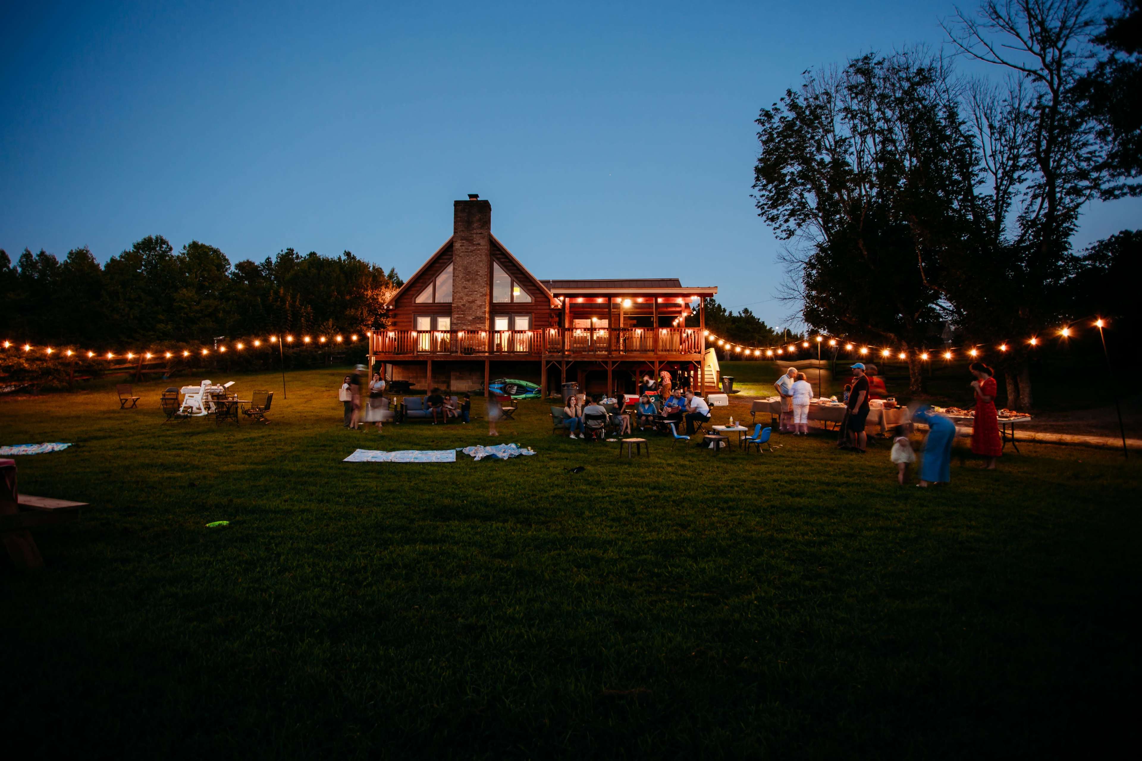 A large house with a deck is illuminated by string lights, surrounded by people gathered on a grassy area in the evening.