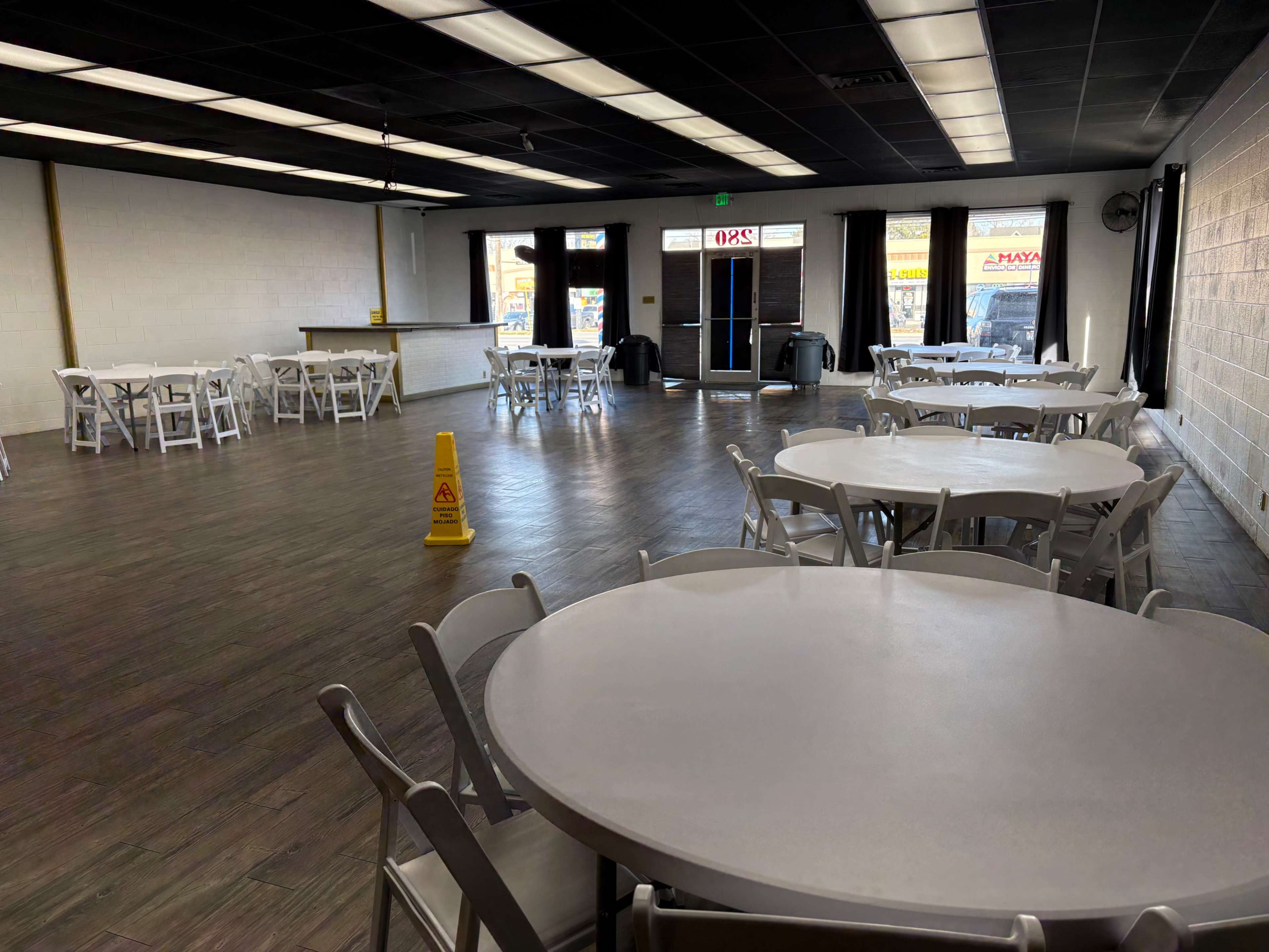 The image shows an empty banquet hall with several round tables and chairs arranged neatly, a caution sign on the floor, and large windows at the front.