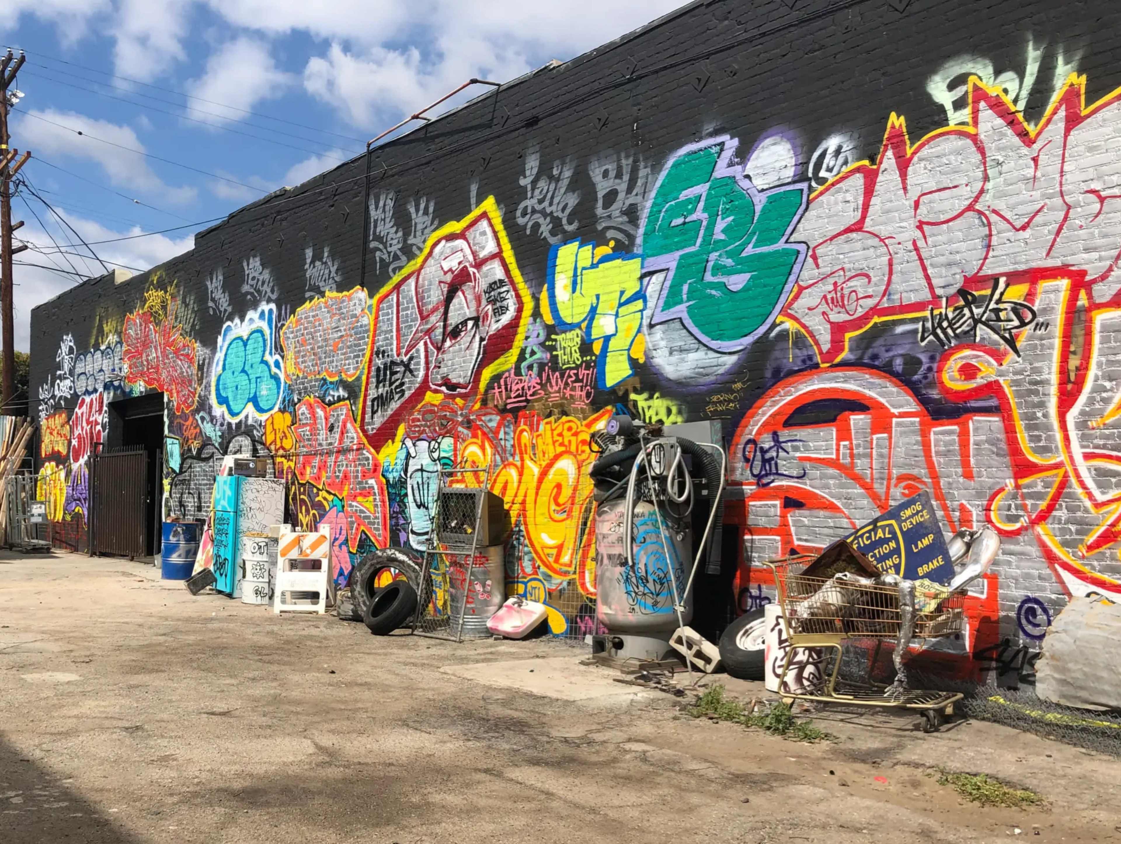 The image shows a graffiti-covered wall in an alleyway, with various colorful tags and artwork alongside discarded shopping carts and road signs.