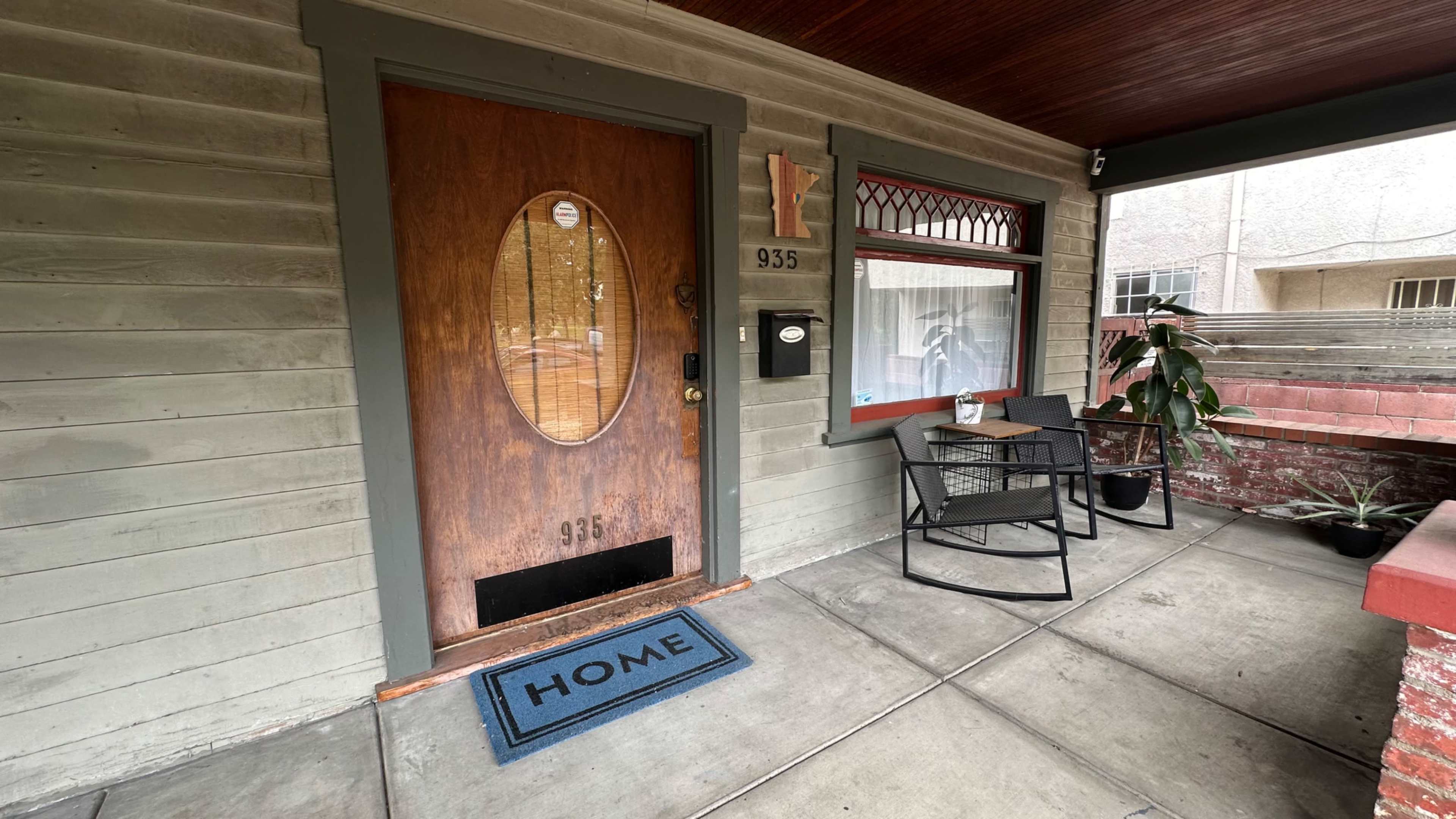 The image shows the entrance of a house with a wooden door featuring an oval window, a welcome mat that reads "HOME," and a small seating area with a chair and potted plant on a covered porch.