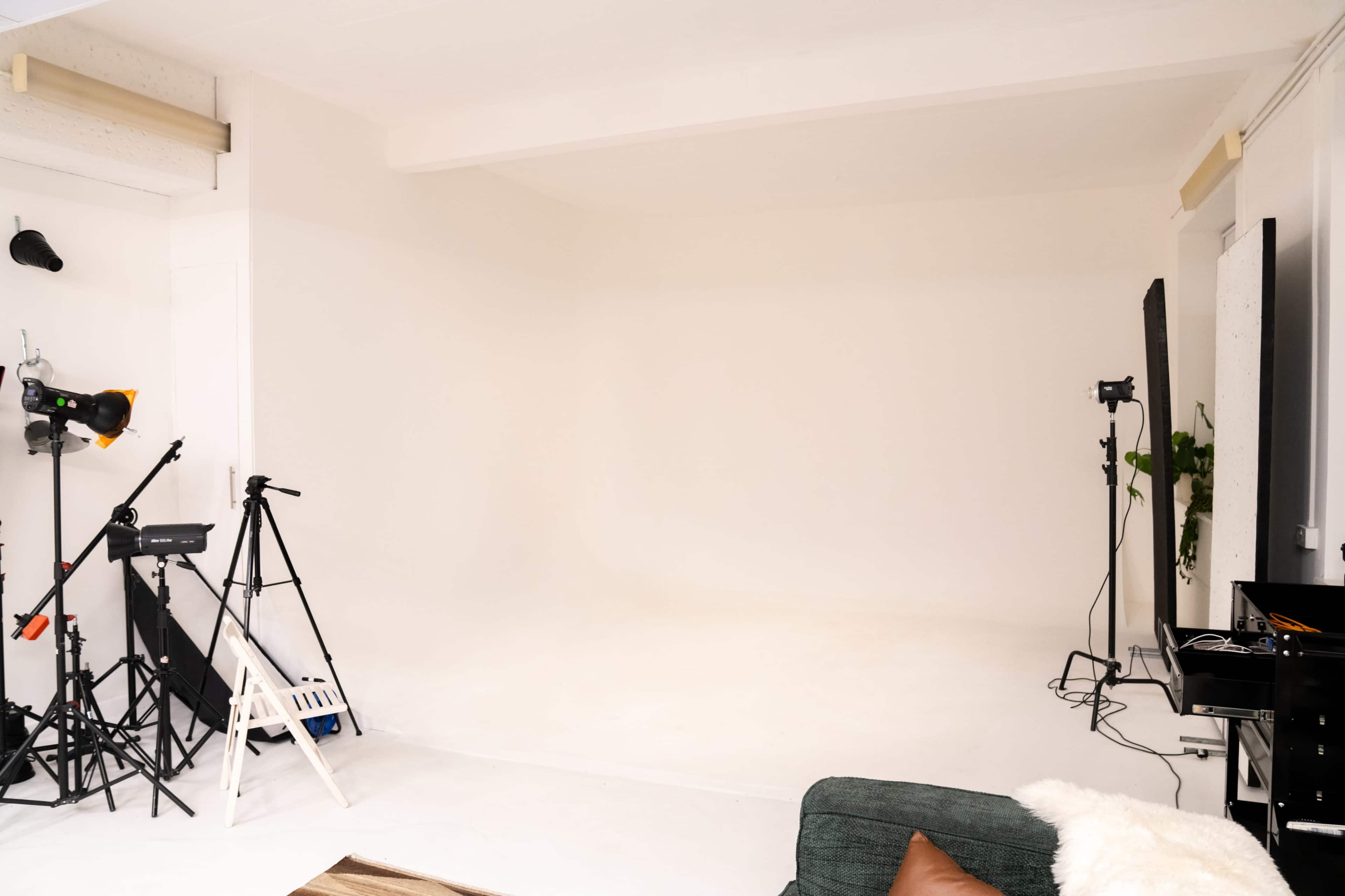 The image shows an empty photo studio with a plain white backdrop and various photography equipment including lights and tripods.