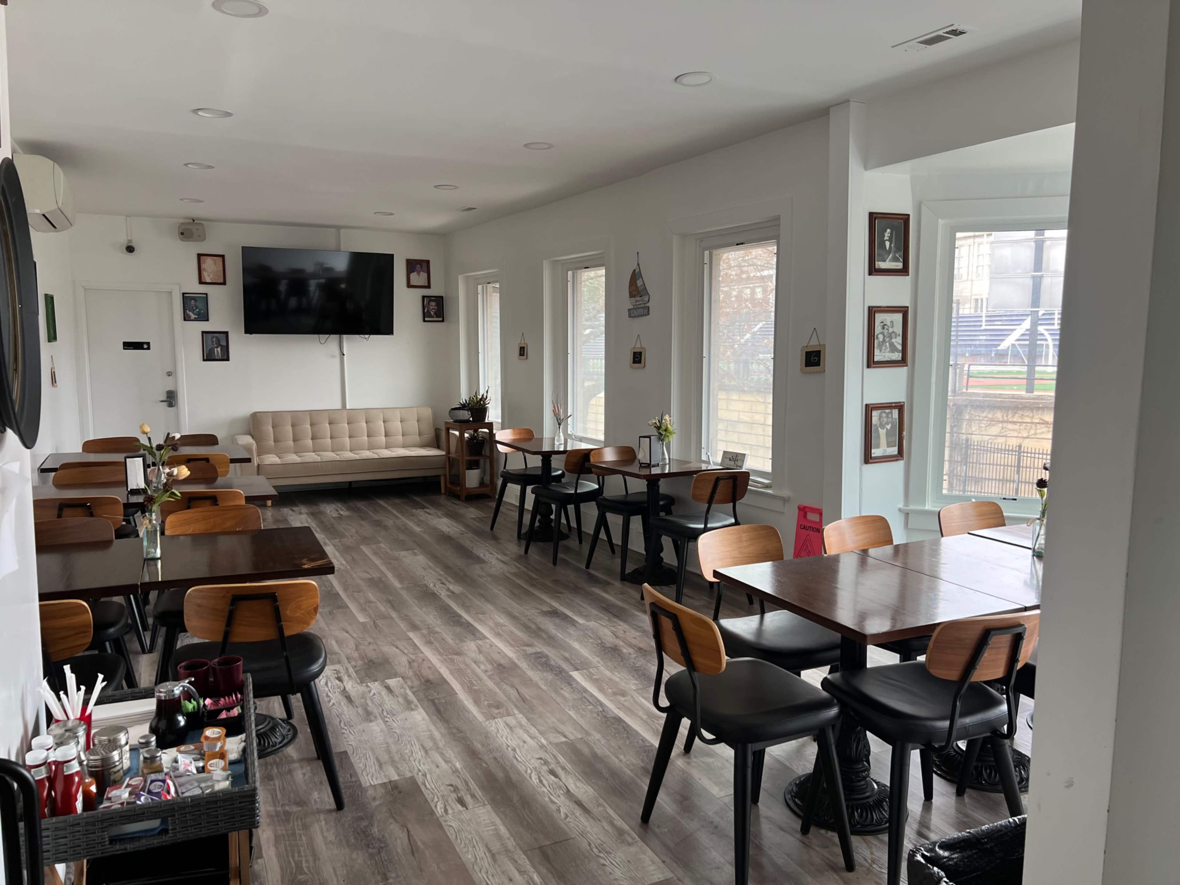 The image shows a spacious cafe with wooden tables and chairs, a couch along one wall, and large windows letting in natural light.