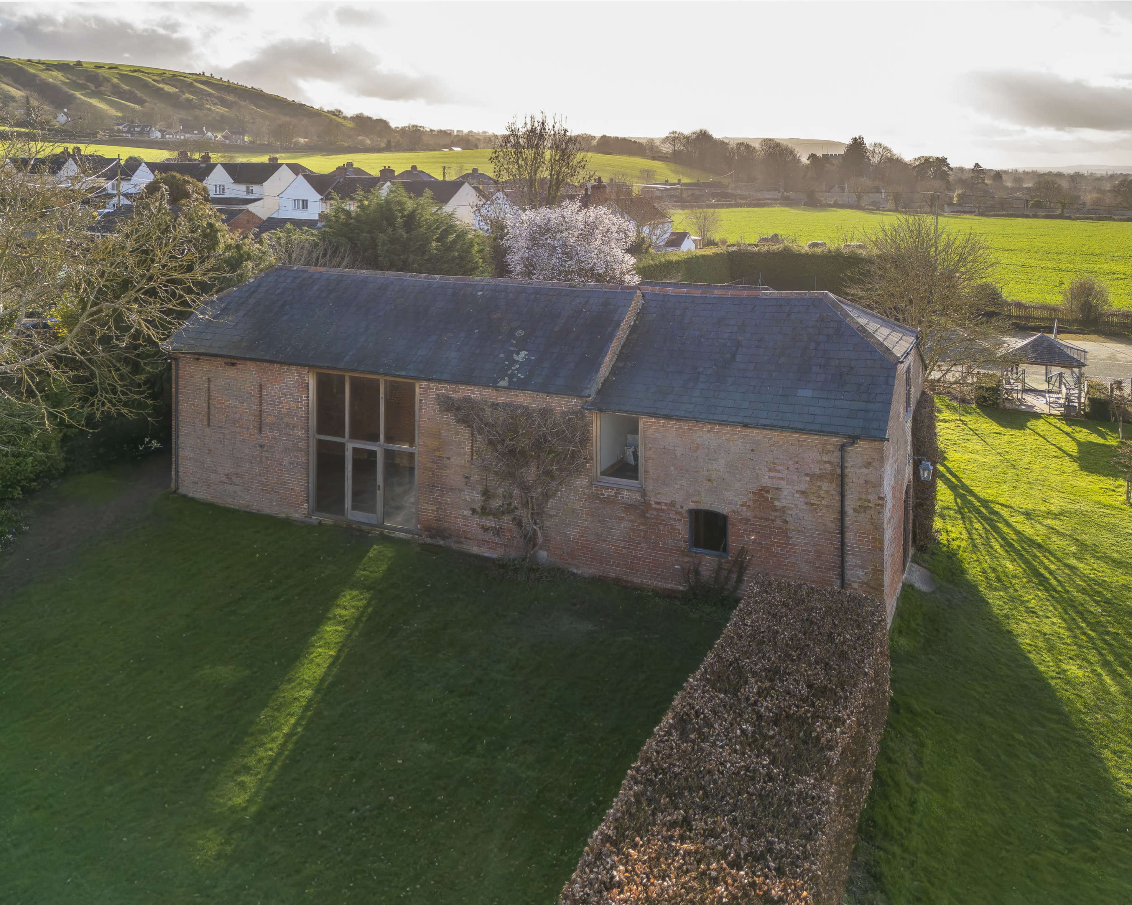 The image shows a brick building with a sloped roof surrounded by green grass and trees, located near rolling hills and houses in the background.