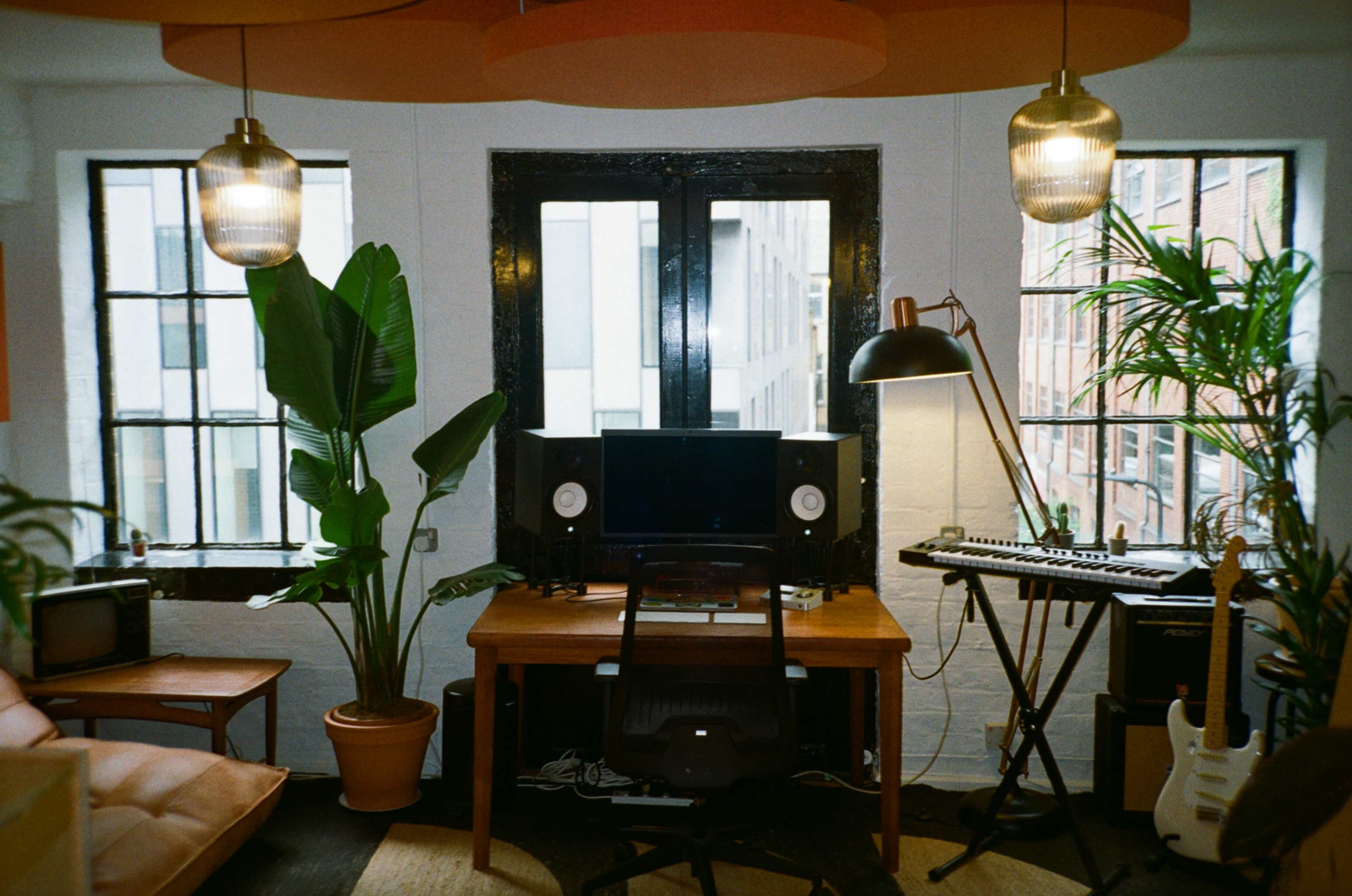 A modern workspace features a desk with a computer and speakers, surrounded by large windows, plants, a guitar, and a keyboard.