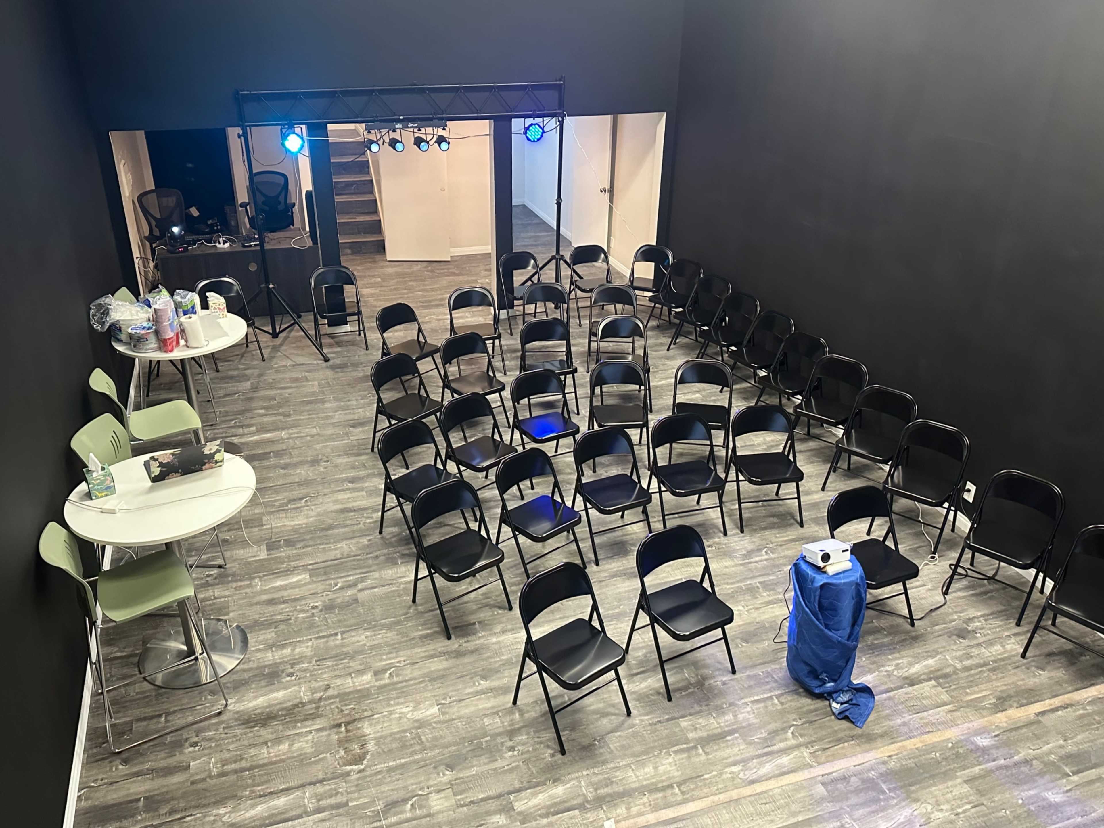 A vacant room arranged with rows of black chairs facing a stage, illuminated by blue lights, with tables and supplies in the background.