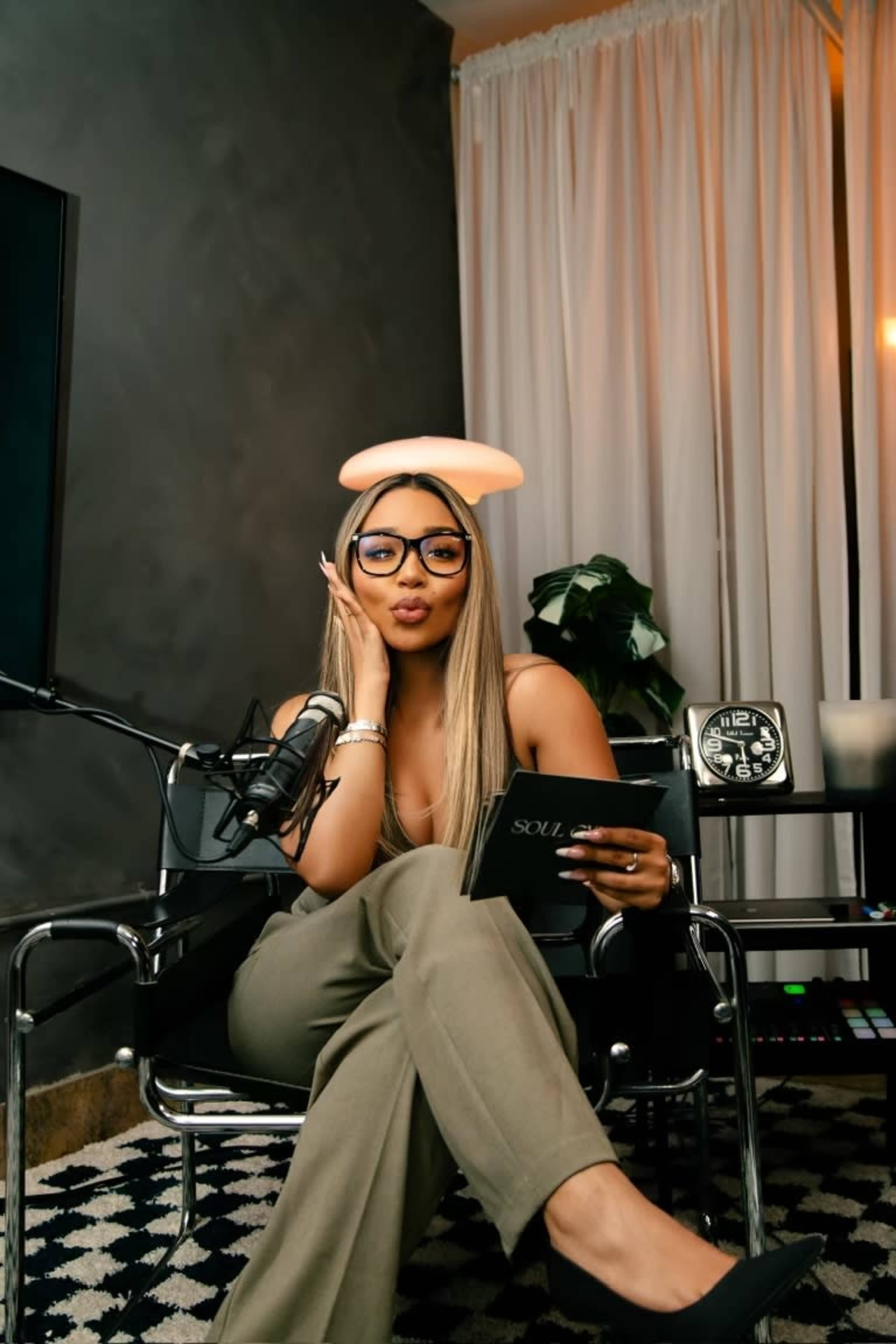 A woman with long hair and glasses sits in a modern chair holding a book, with a microphone and a decorative clock nearby in a well-lit room.