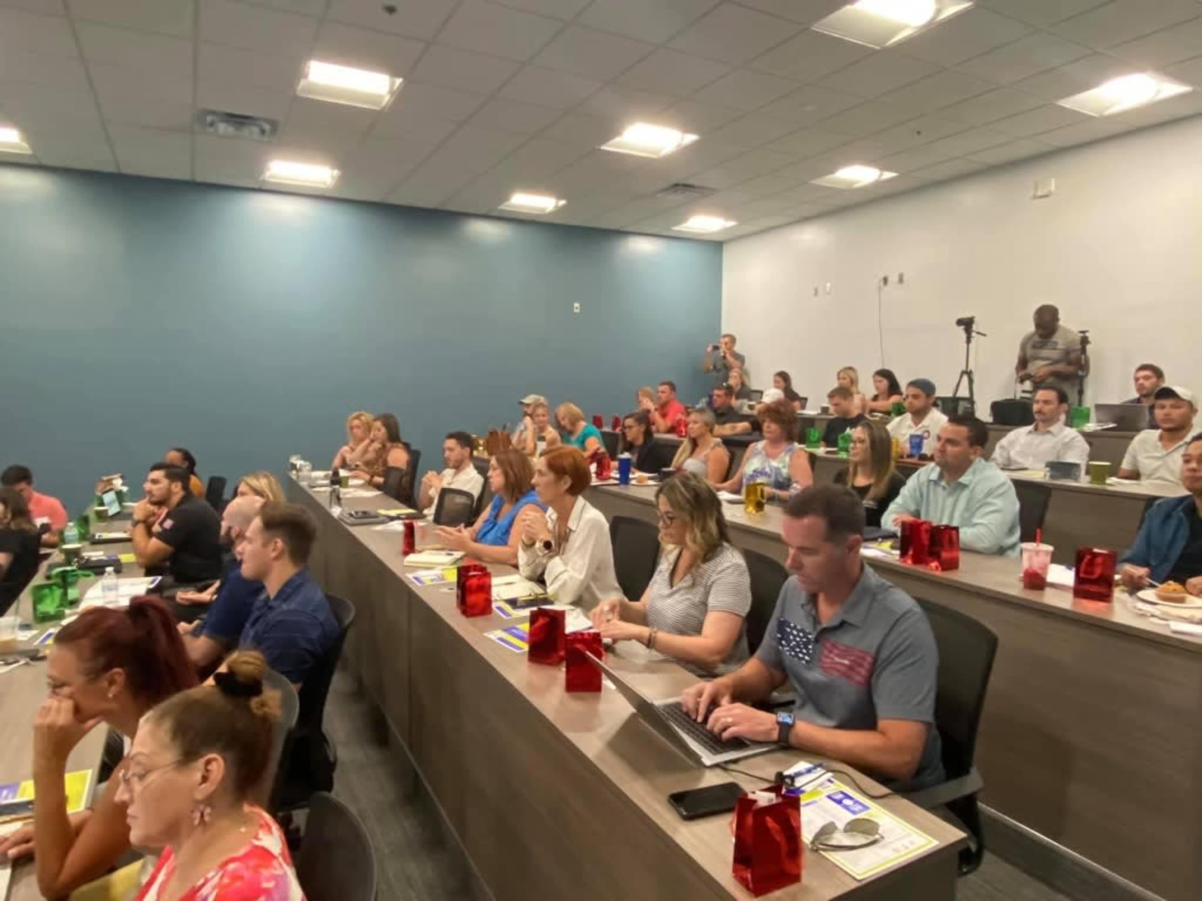 A diverse group of attendees sits in a classroom-style setting, focused on a presentation with notebooks and laptops in front of them.