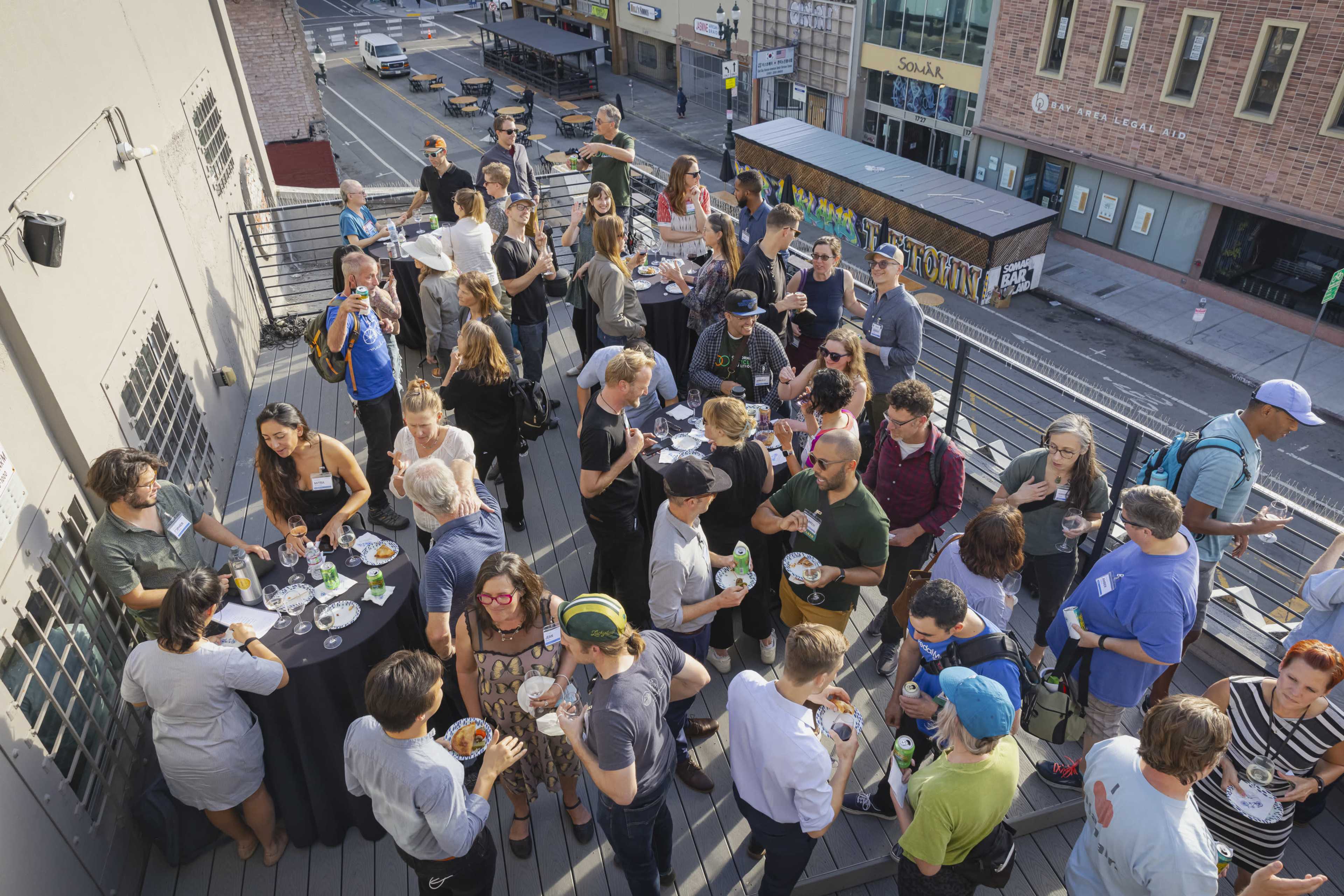A large group of people socializes and enjoys food and drinks on a rooftop deck overlooking a city street.
