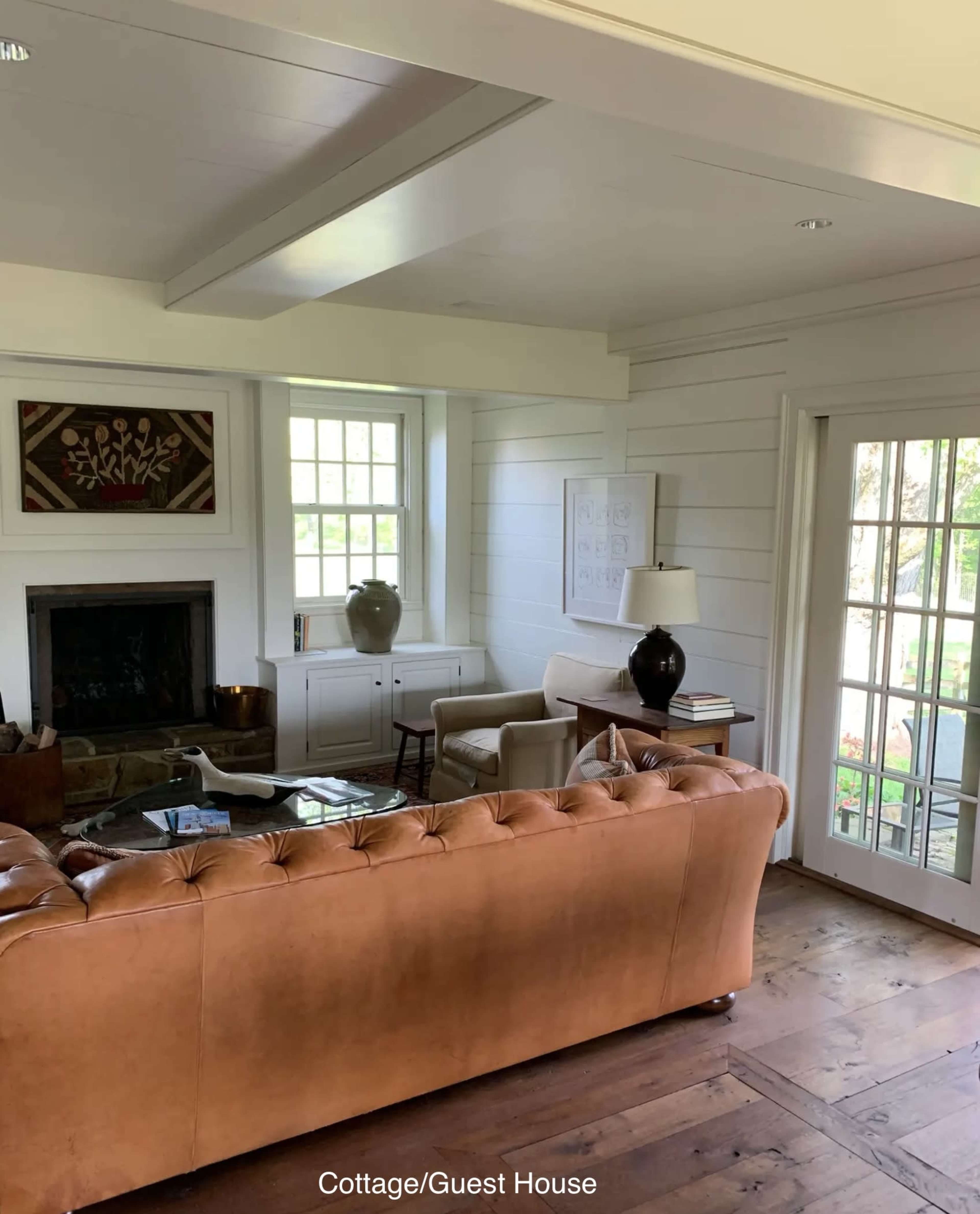 The image shows a cozy living room with a brown tufted leather sofa, a fireplace, and large windows that open to a garden.
