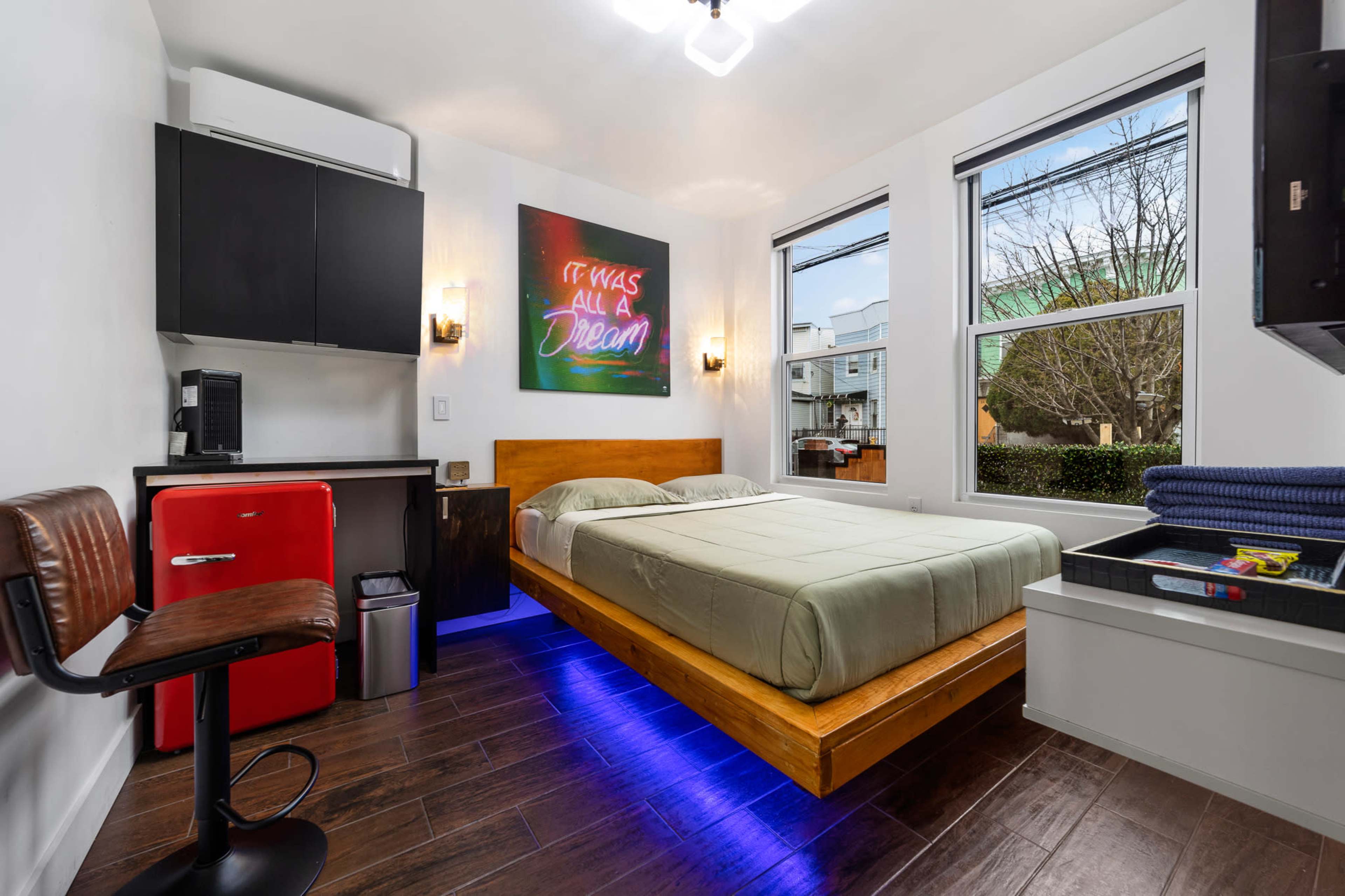 The image shows a modern bedroom featuring a floating bed, a compact kitchenette with a refrigerator, and large windows allowing natural light.