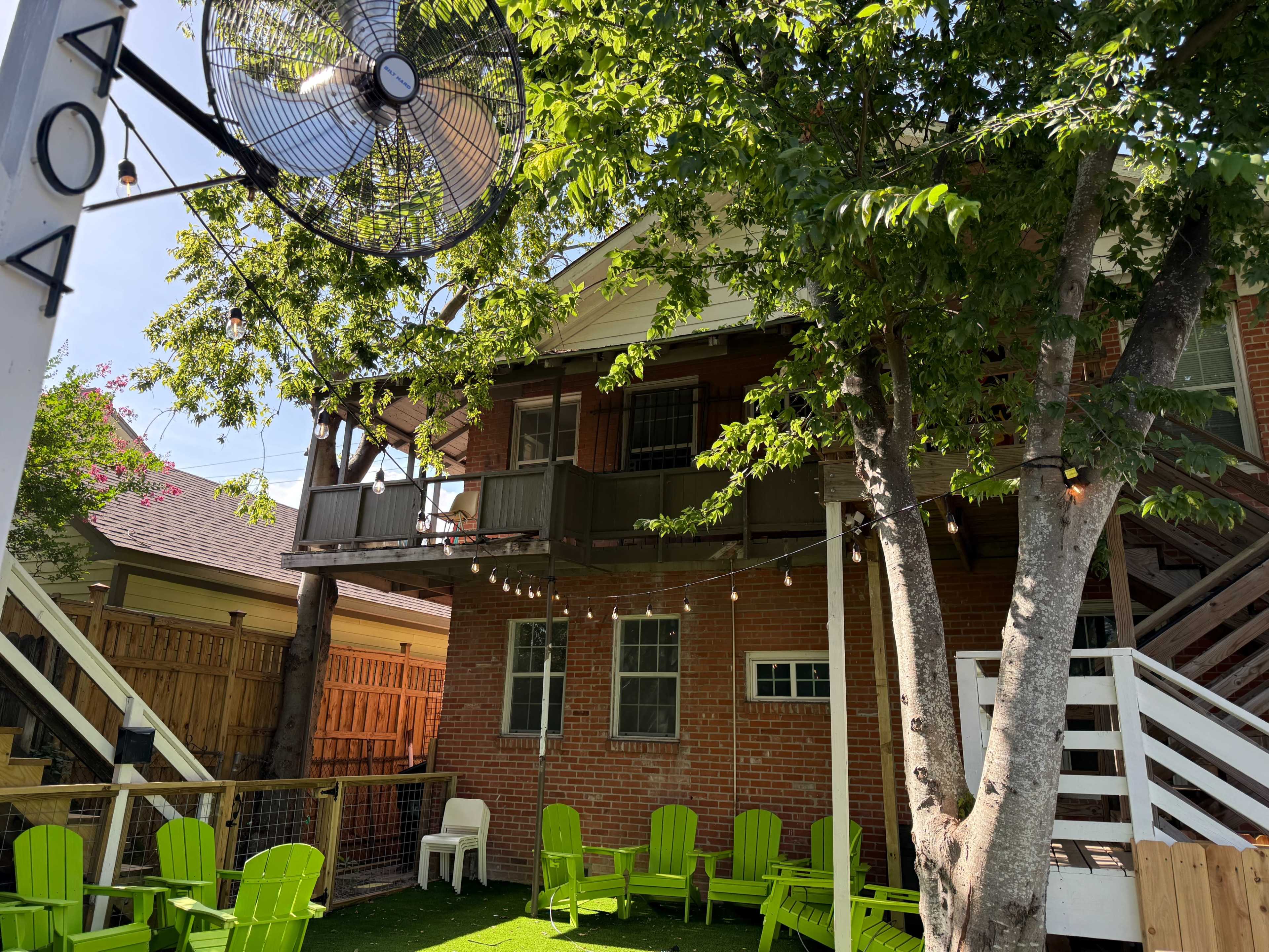 The image shows a backyard space with green chairs, a large outdoor fan, and a two-story brick building surrounded by trees and string lights.