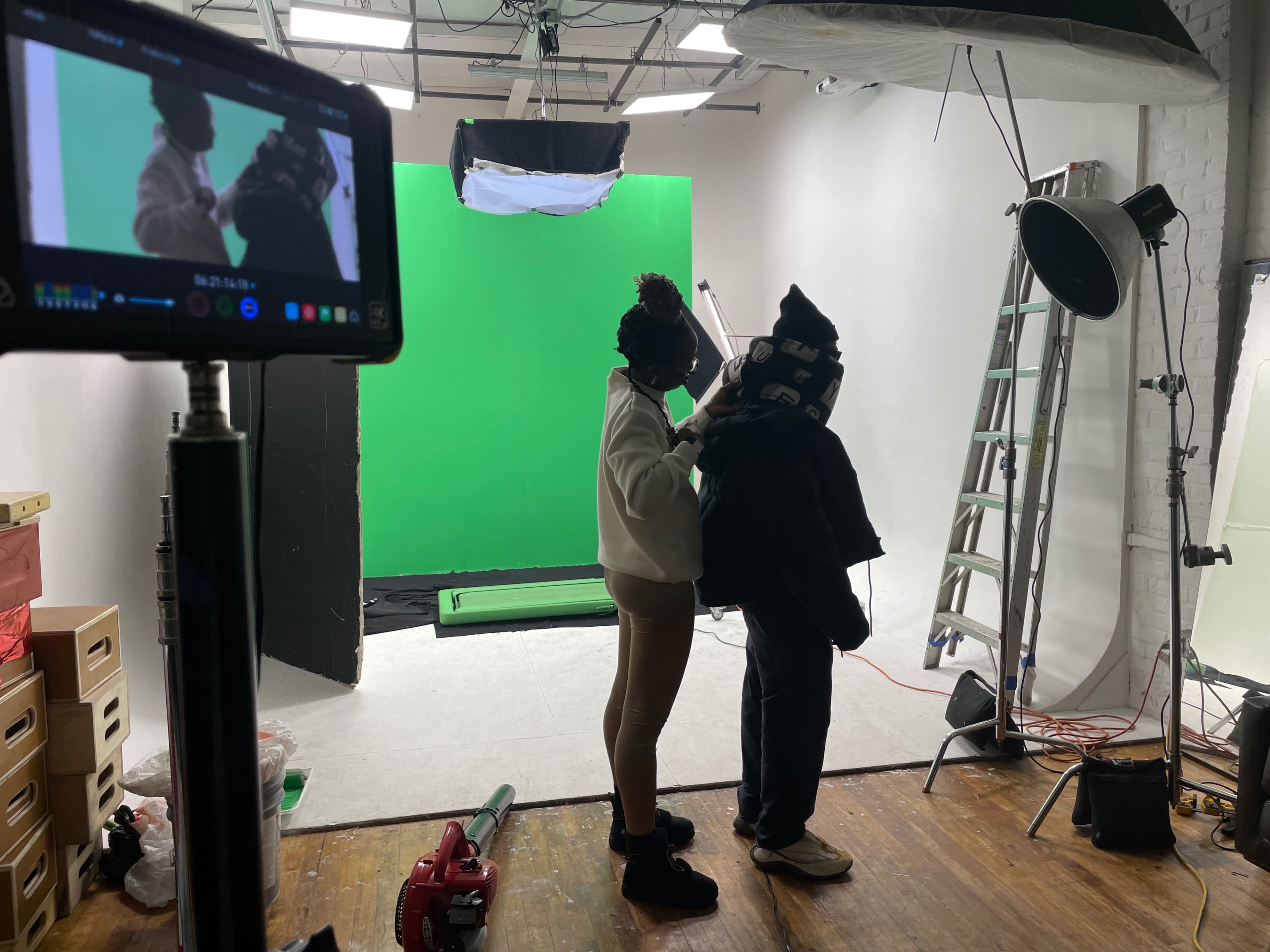 A person is adjusting clothing on another individual in a studio setting with a green screen and various lighting equipment.