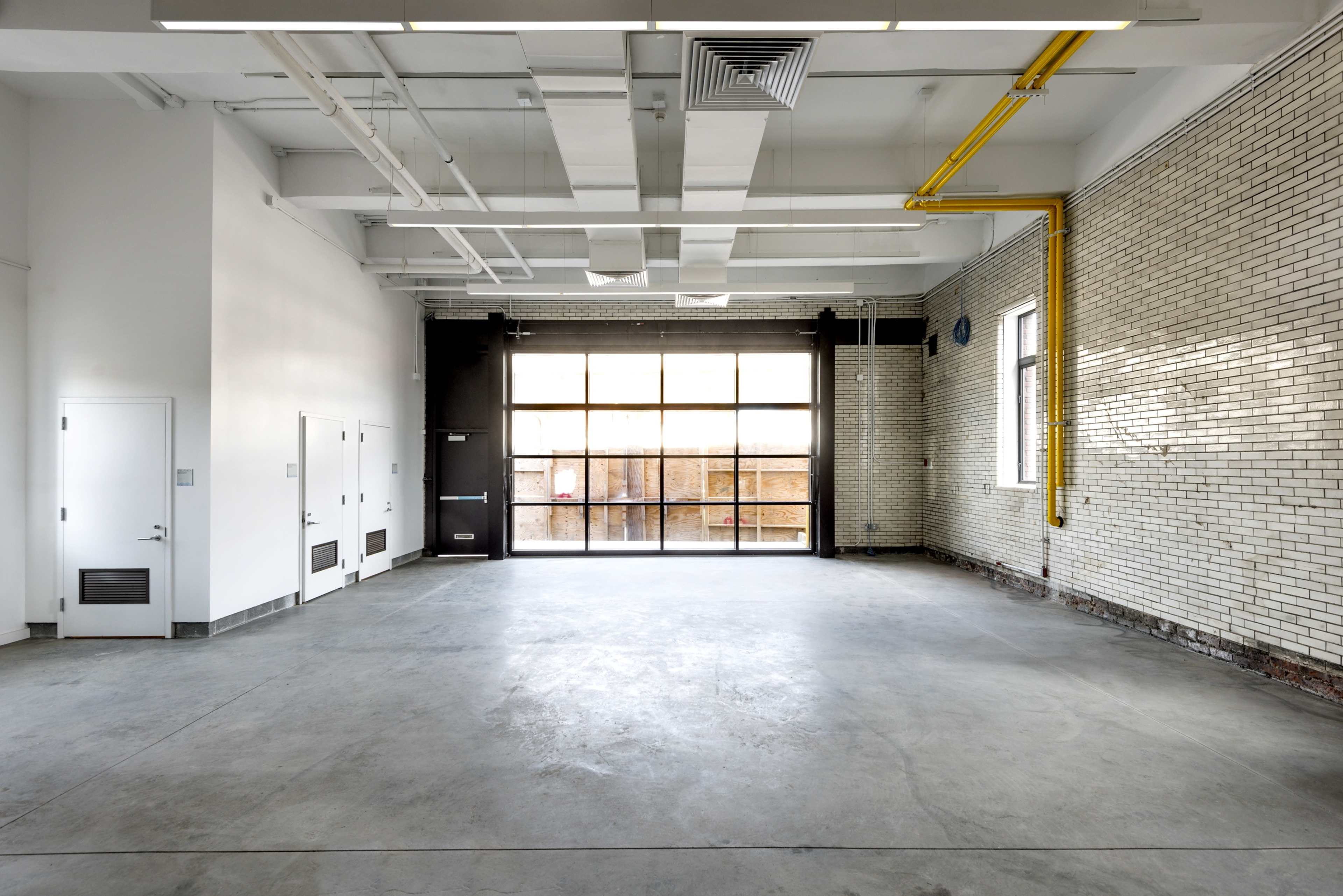 The interior of a spacious, empty room features white brick walls, a concrete floor, and large windows that provide natural light.