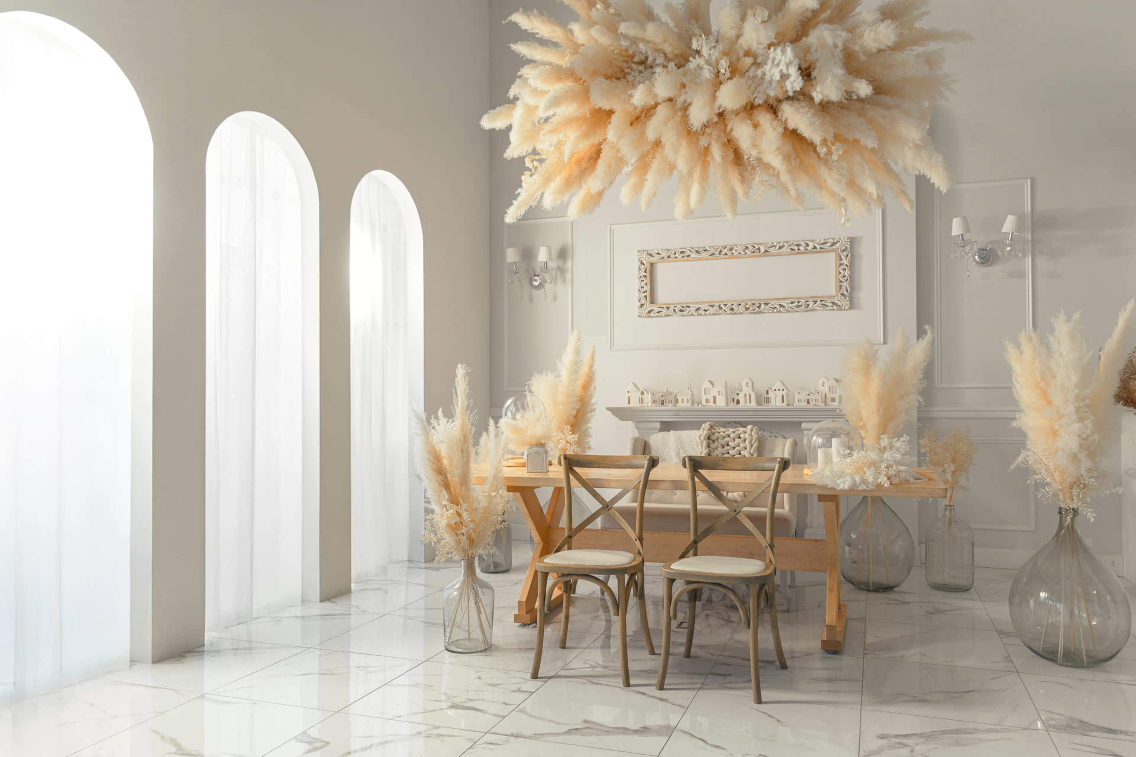 A bright dining room features a wooden table with two chairs, surrounded by large vases filled with pampas grass, and large windows that allow natural light to fill the space.