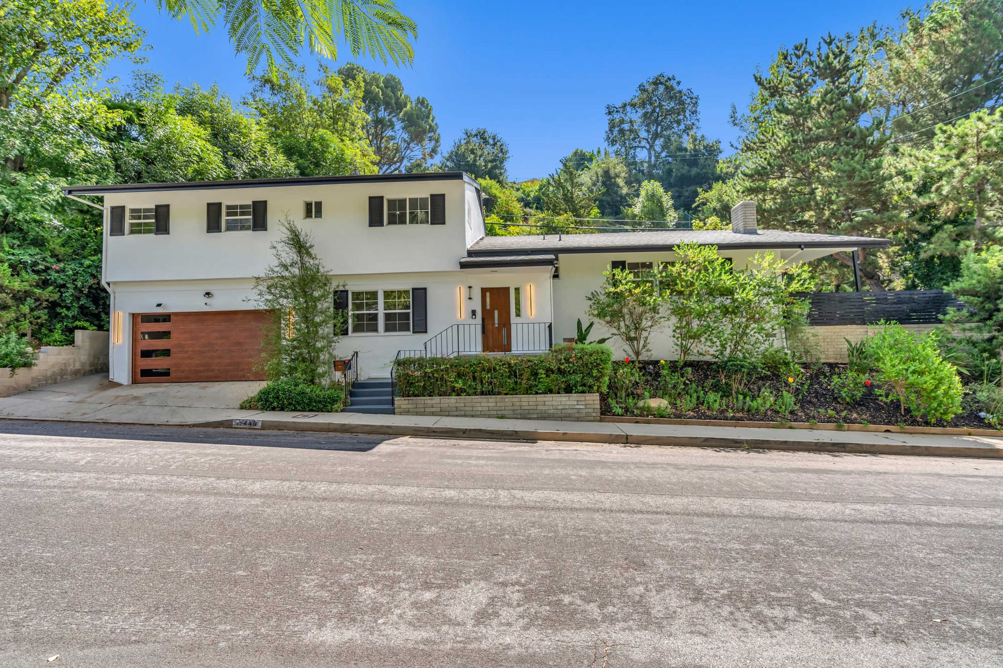 A two-story white house with a brown garage door is surrounded by greenery along a quiet street.