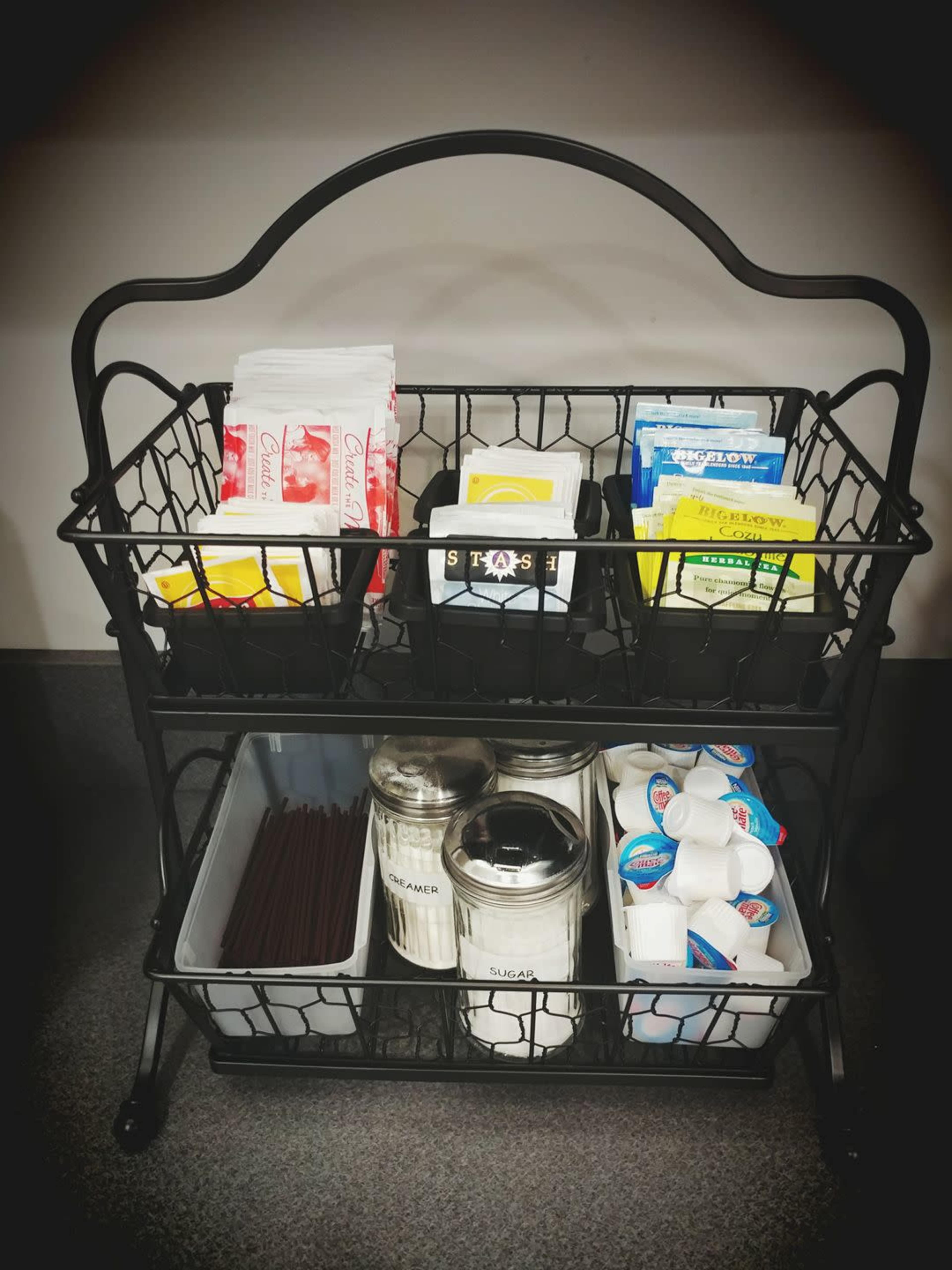 A black wire rack contains various packets of tea, sugar, and creamers on its upper shelf, while the lower shelf holds glass containers for sugar and a box of stirrers.