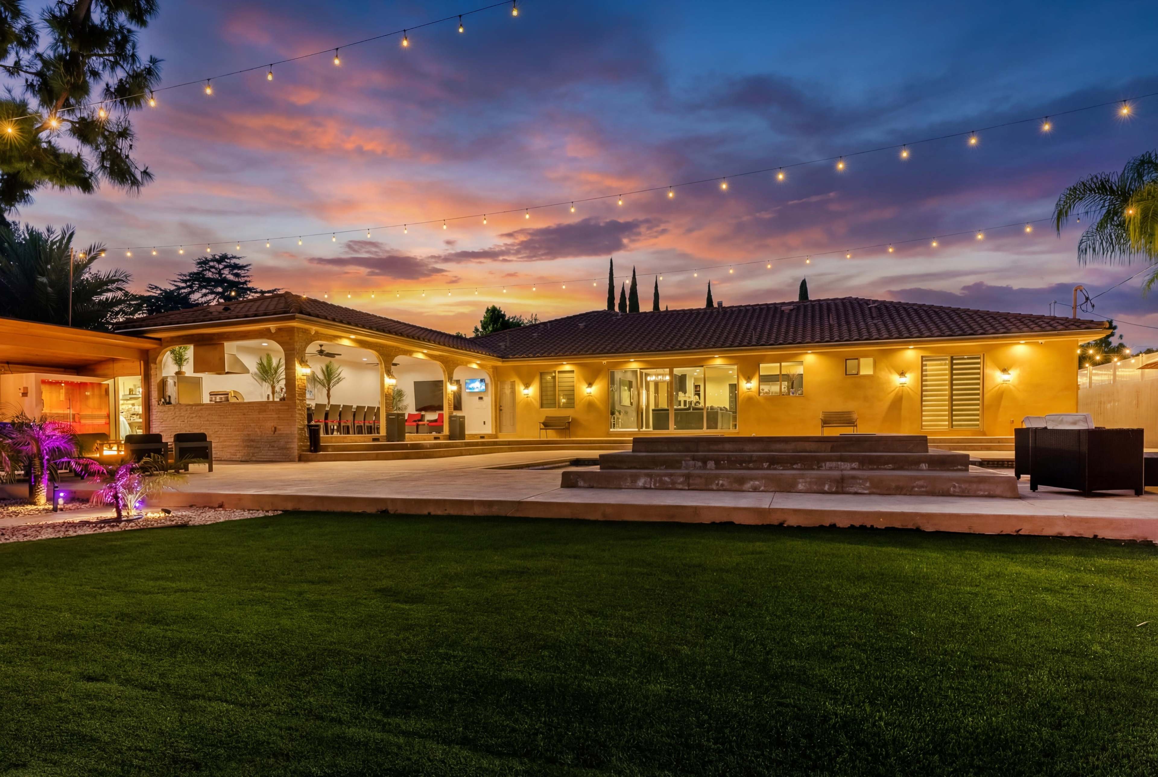 A modern single-story home with a landscaped yard and outdoor lighting, set against a colorful sunset sky.