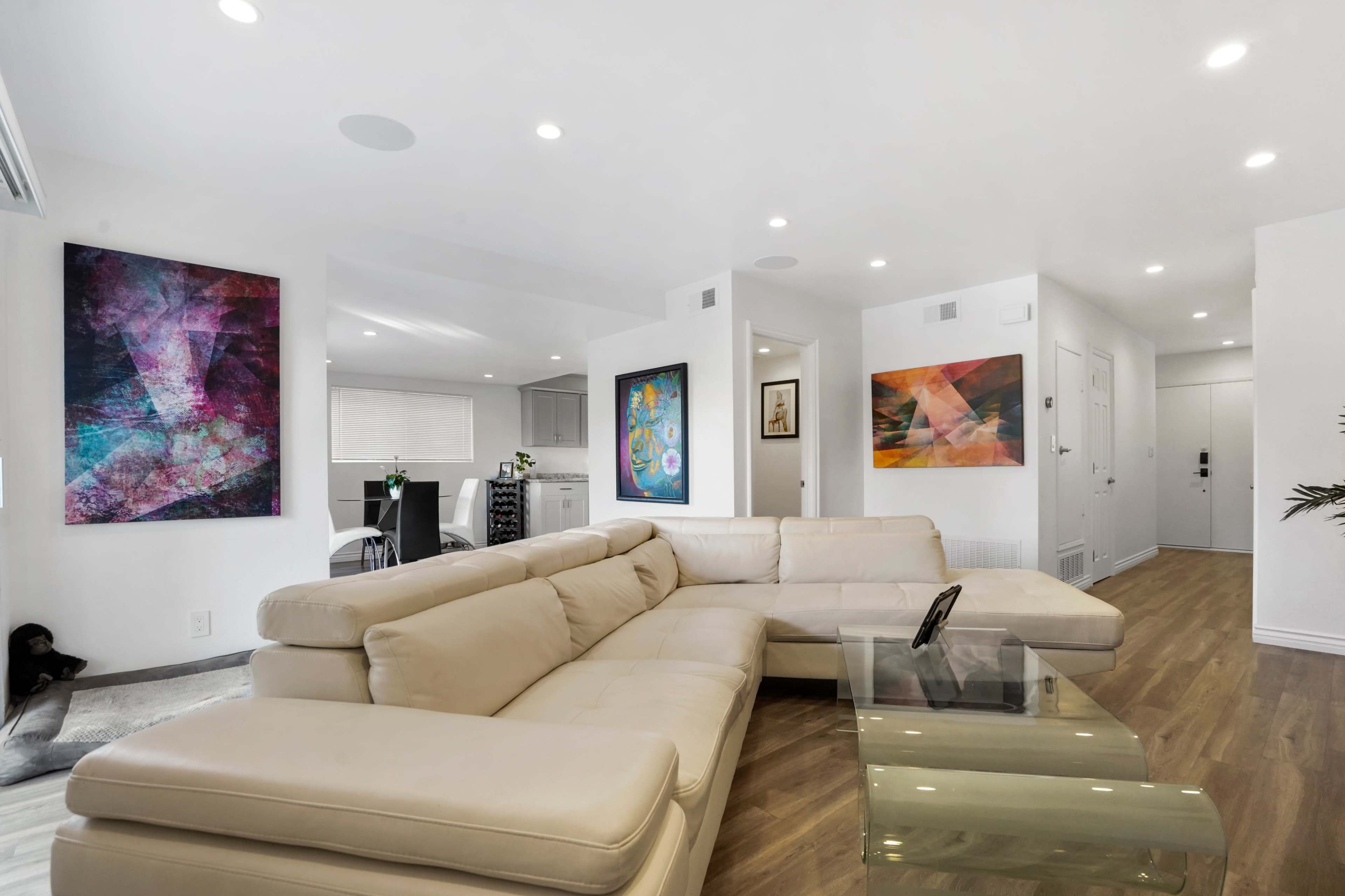 A modern living room with a beige sectional sofa, colorful abstract artwork on the walls, and an open layout leading to a dining area.