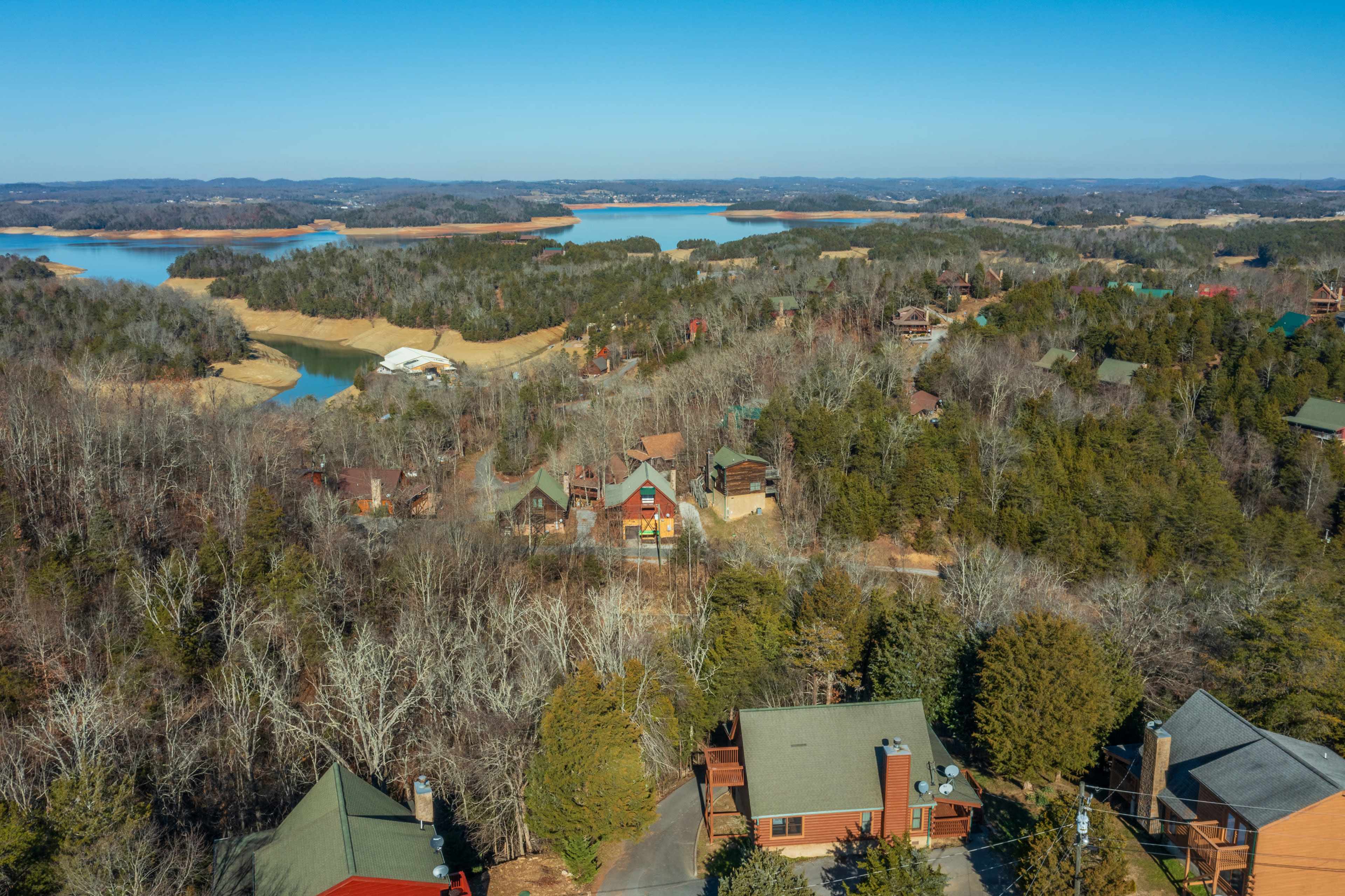 The image shows a scenic view of a wooded area with a cluster of homes and a lake in the background, surrounded by hills.