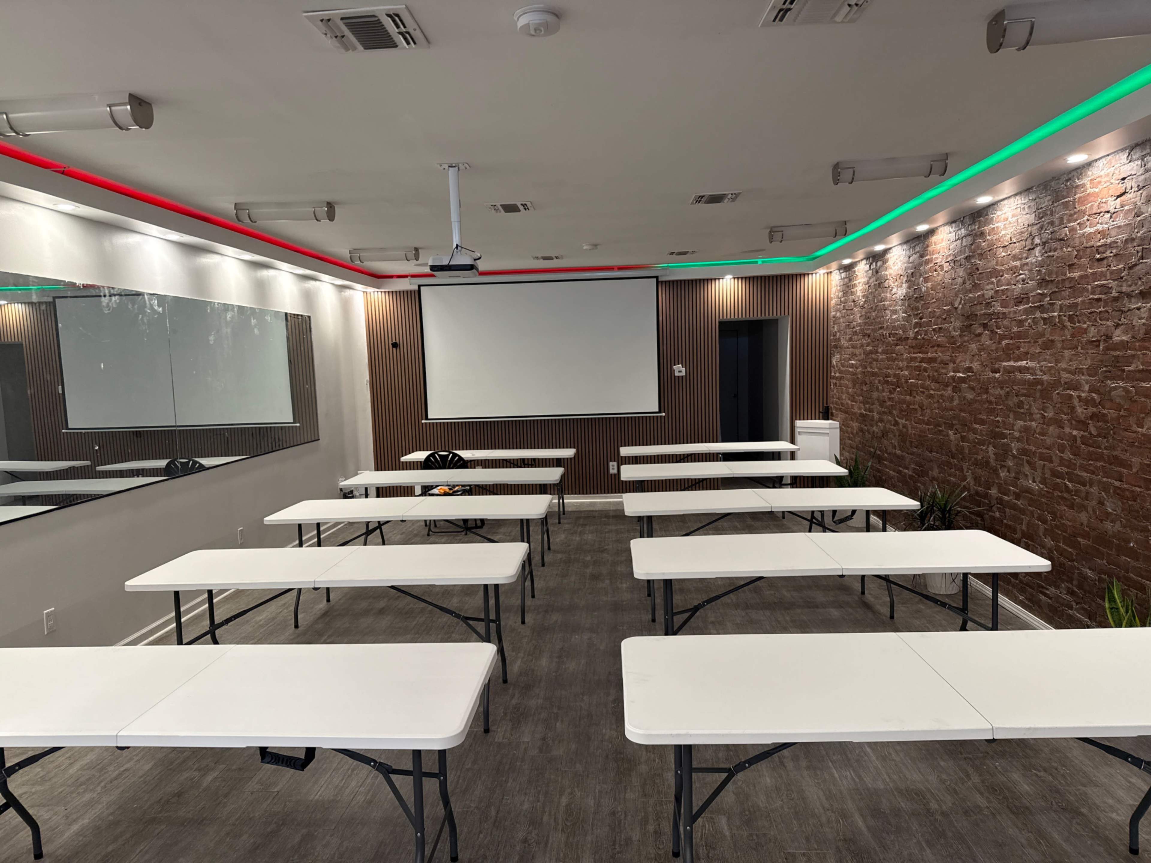 The image shows a classroom with several rows of white folding tables and a large screen at the front, accented by colorful overhead lighting and a brick wall.