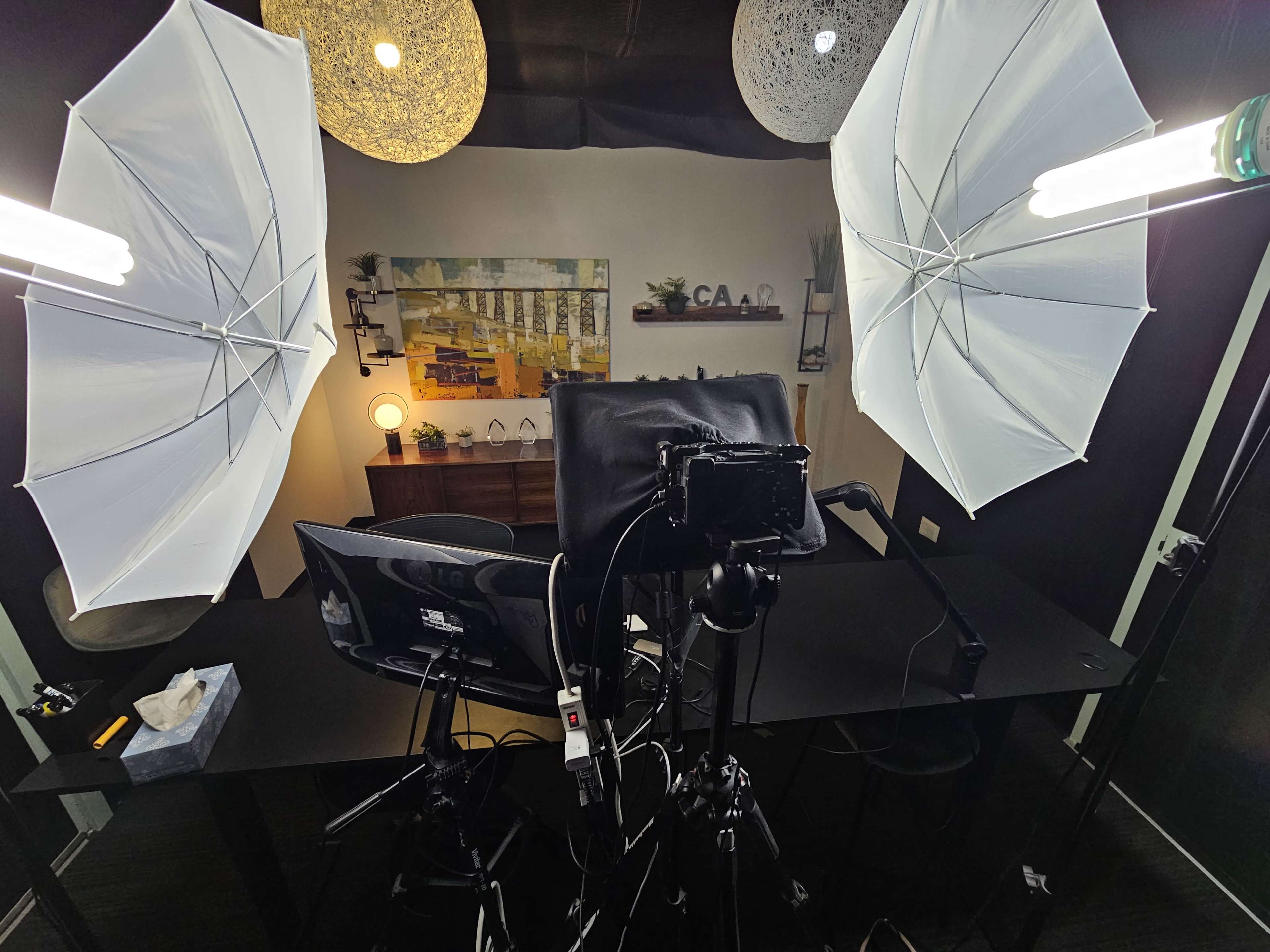 A studio setup with a camera on a tripod facing a desk, surrounded by two softbox lights and a monitor.
