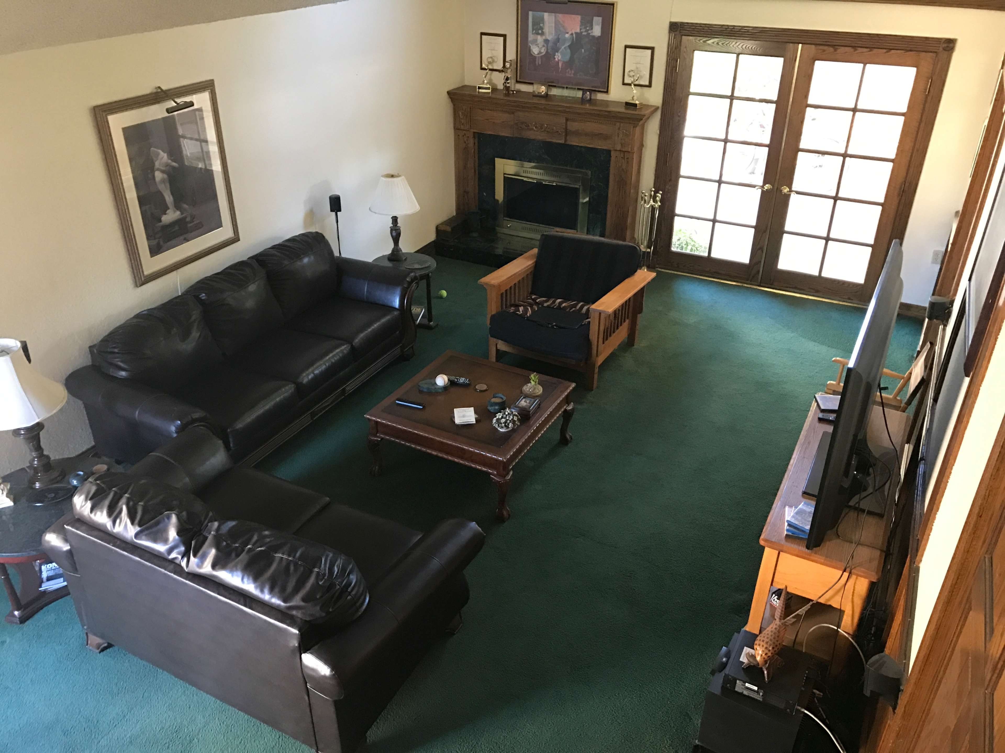 The image shows a living room with two black leather sofas, a wooden coffee table, a television, and a fireplace, alongside large patio doors leading outside.