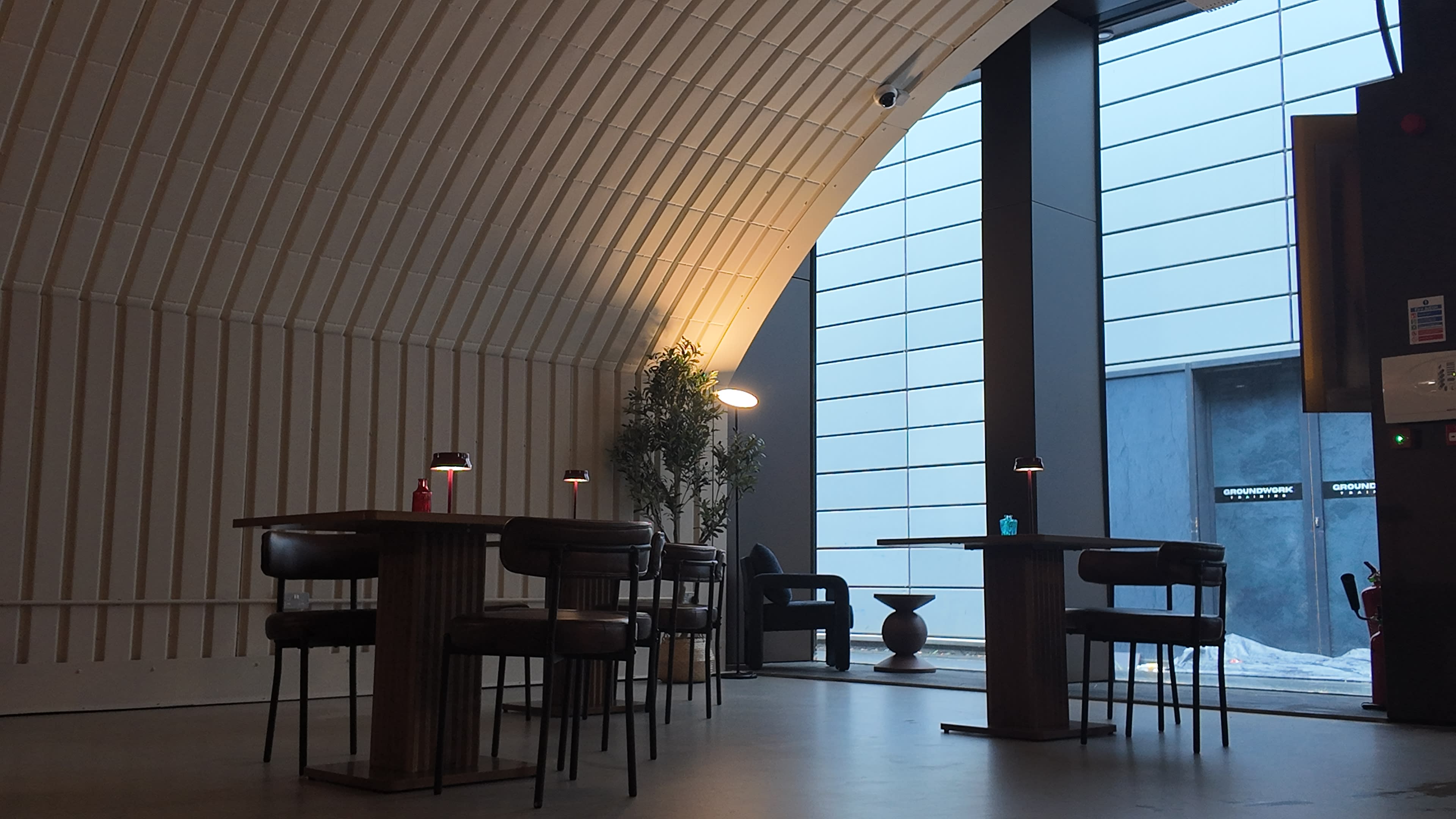 The image shows a modern interior of a café with wooden tables and chairs, featuring large windows that provide natural light.