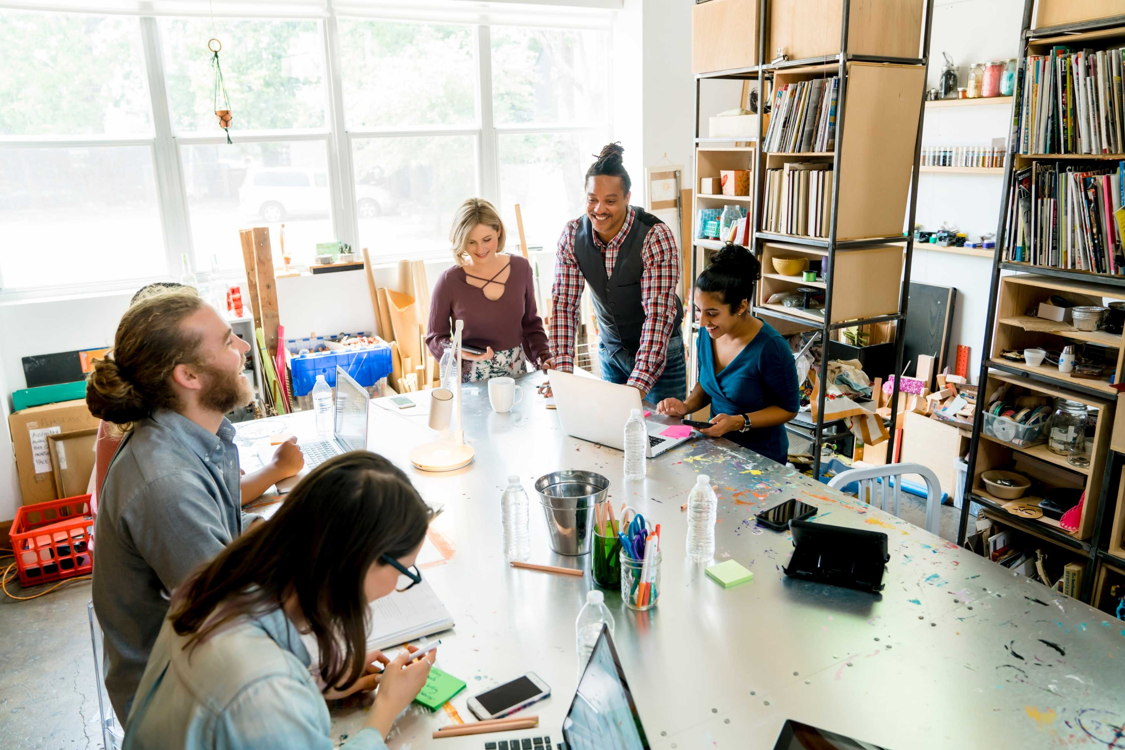 A group of six people collaborates around a large table filled with notebooks and laptops in a bright, well-organized studio.