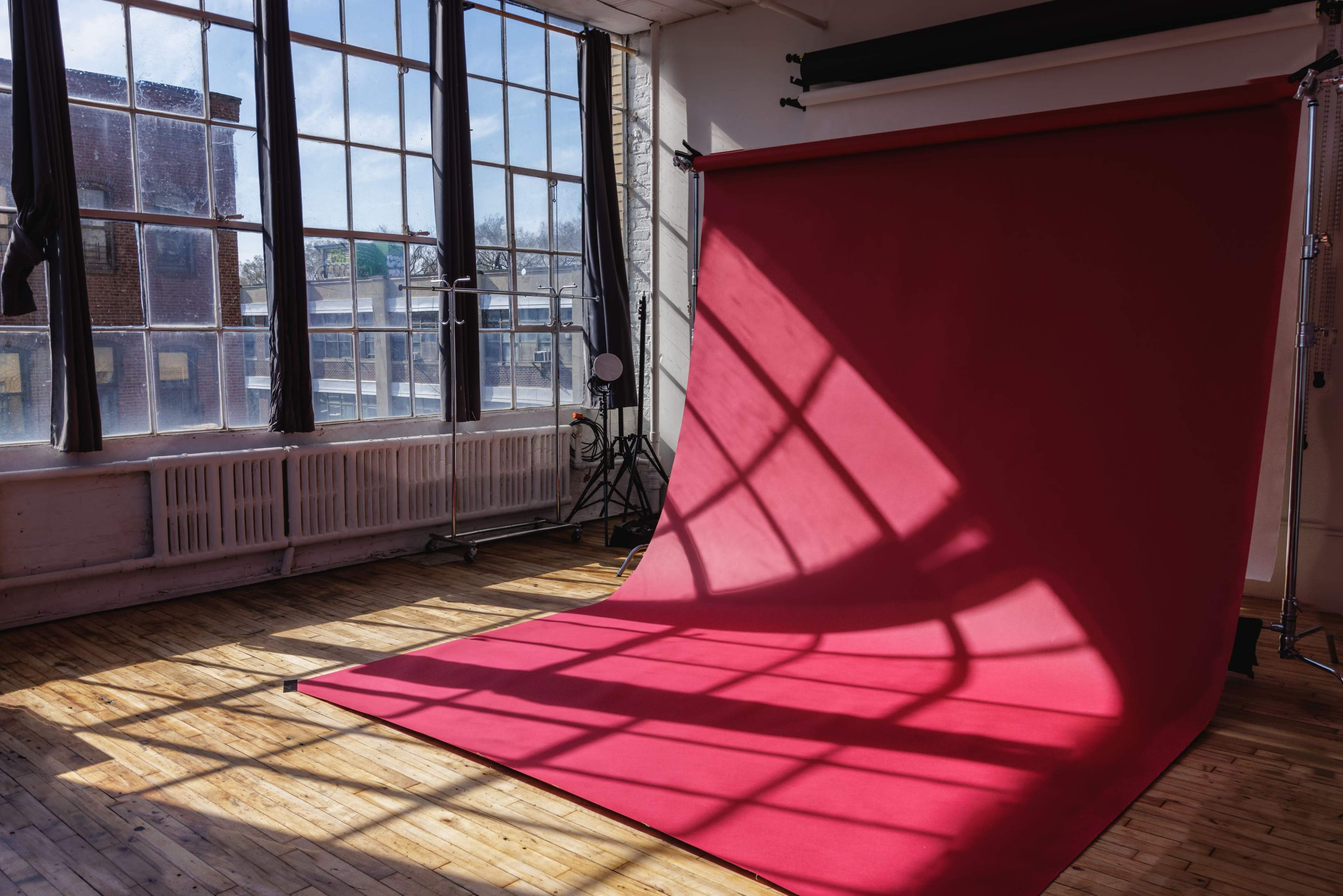 A photography studio features a large red backdrop with sunlight casting shadows across the wooden floor.
