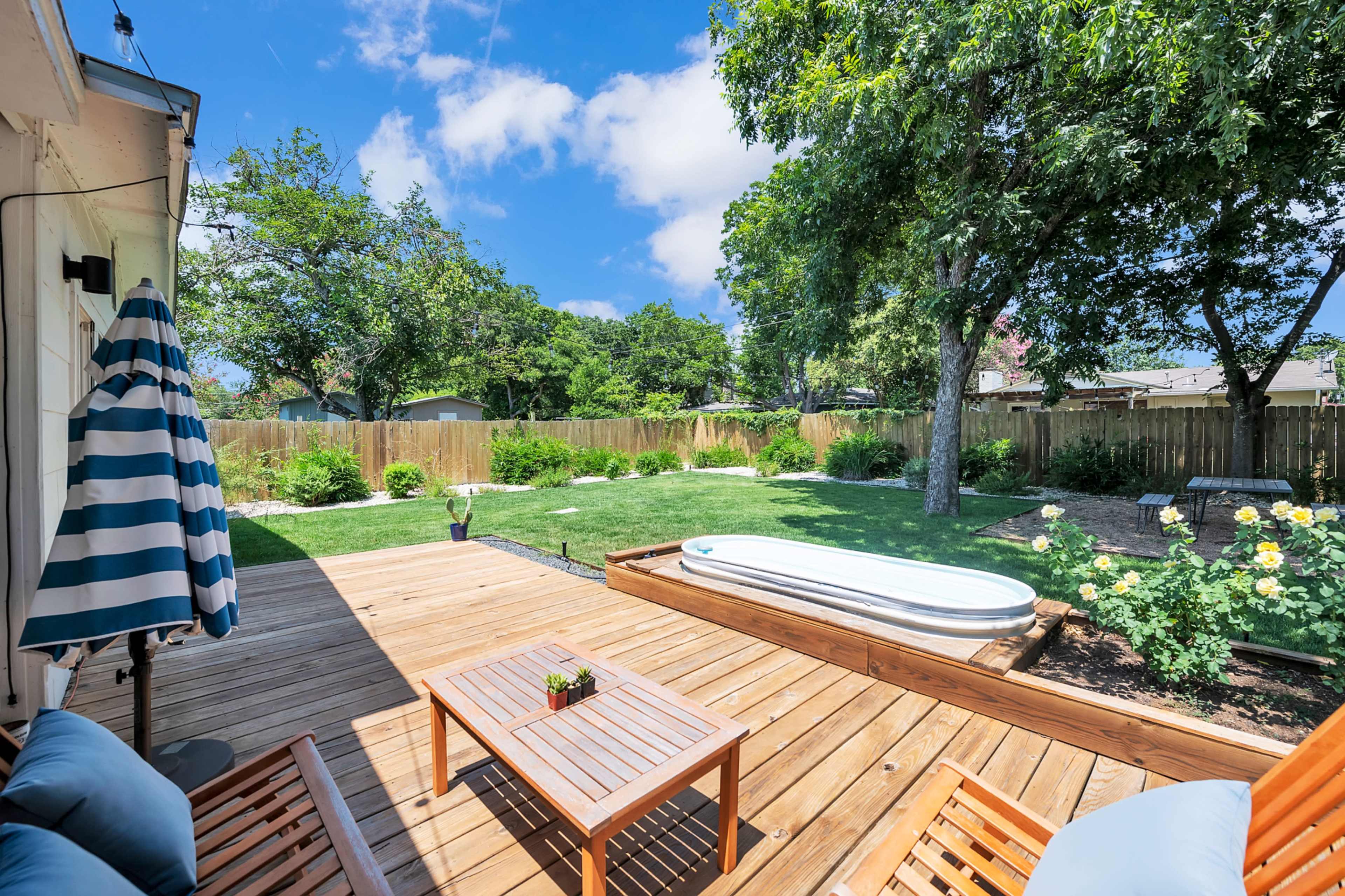 The image shows a backyard deck with wooden seating, a small table, and a white tub, surrounded by green grass and trees under a partly cloudy sky.