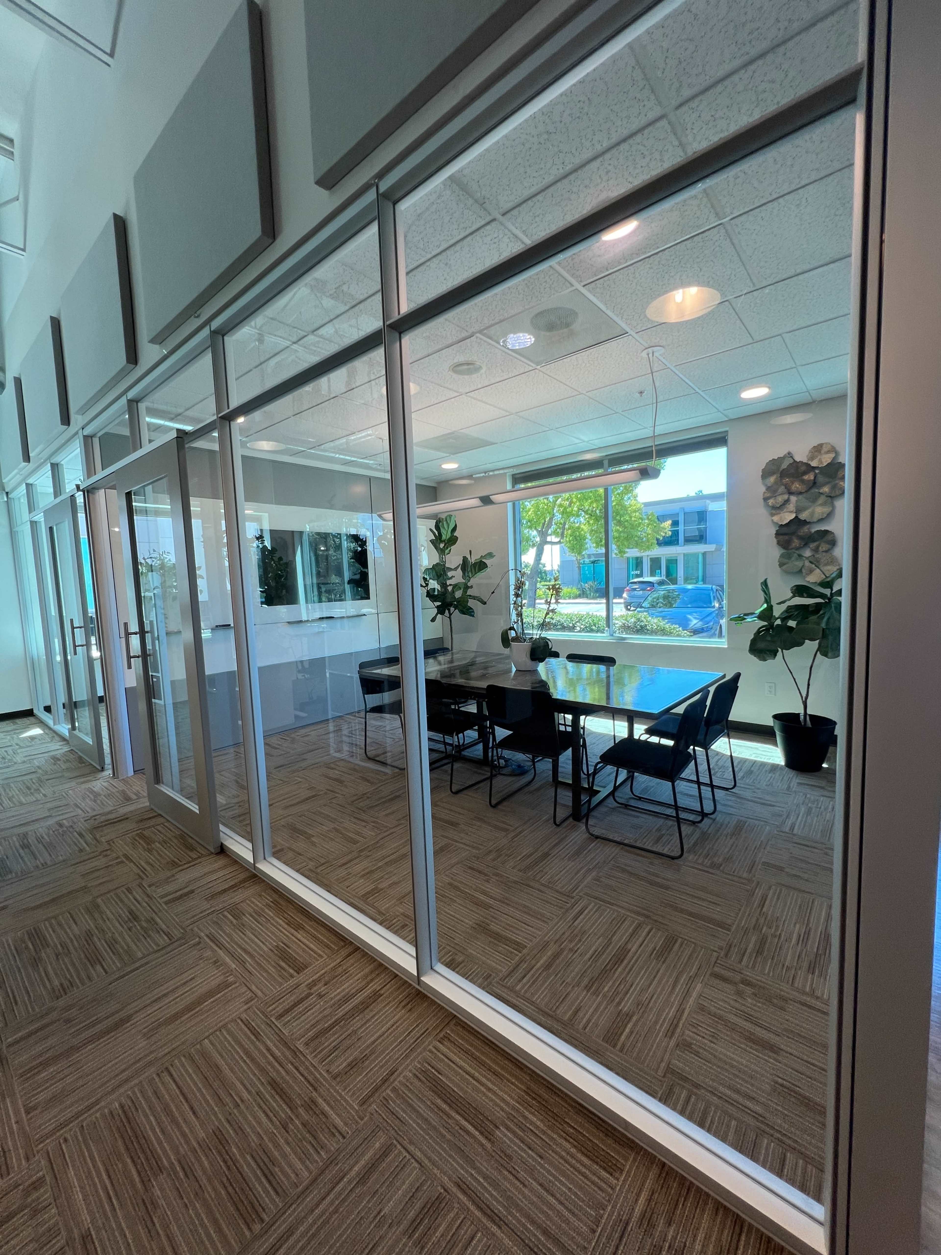 A glass-walled conference room features a table and chairs, surrounded by plants, with natural light streaming in from large windows.