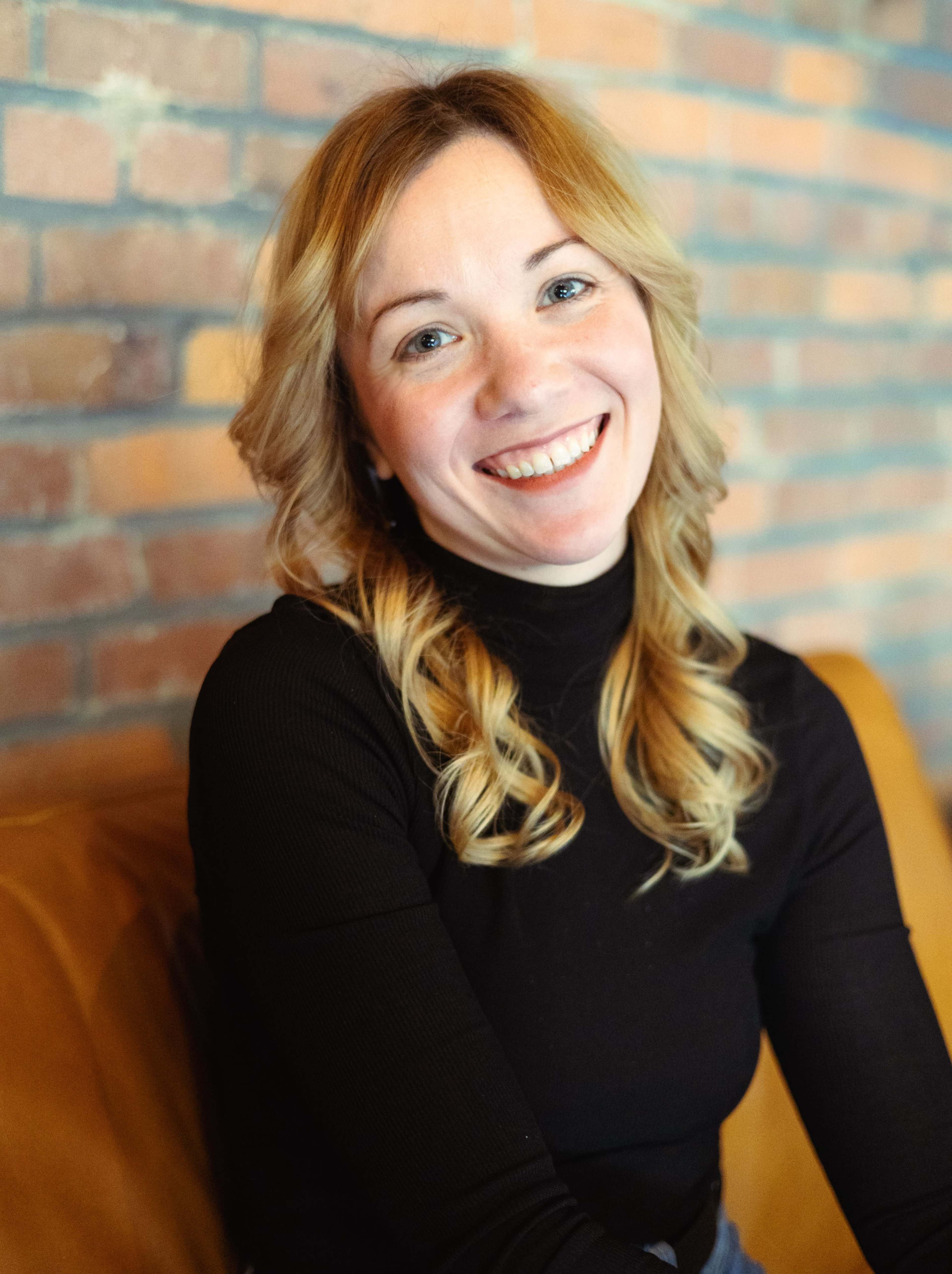A woman with long, wavy hair is smiling while seated against a brick wall.