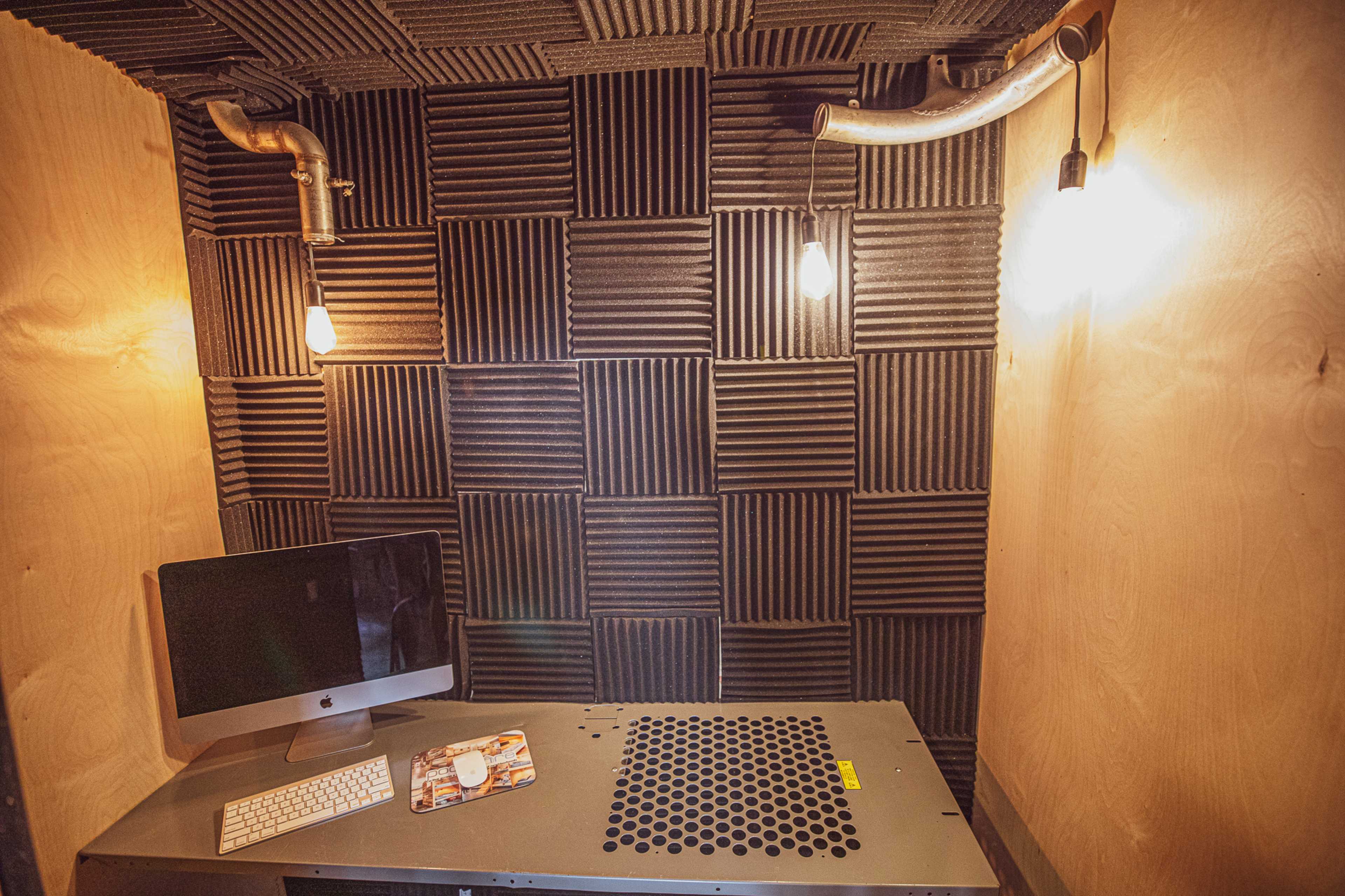 The image shows a small soundproof room featuring a desk with an Apple computer, two exposed light bulbs, and acoustic foam paneling on the walls.