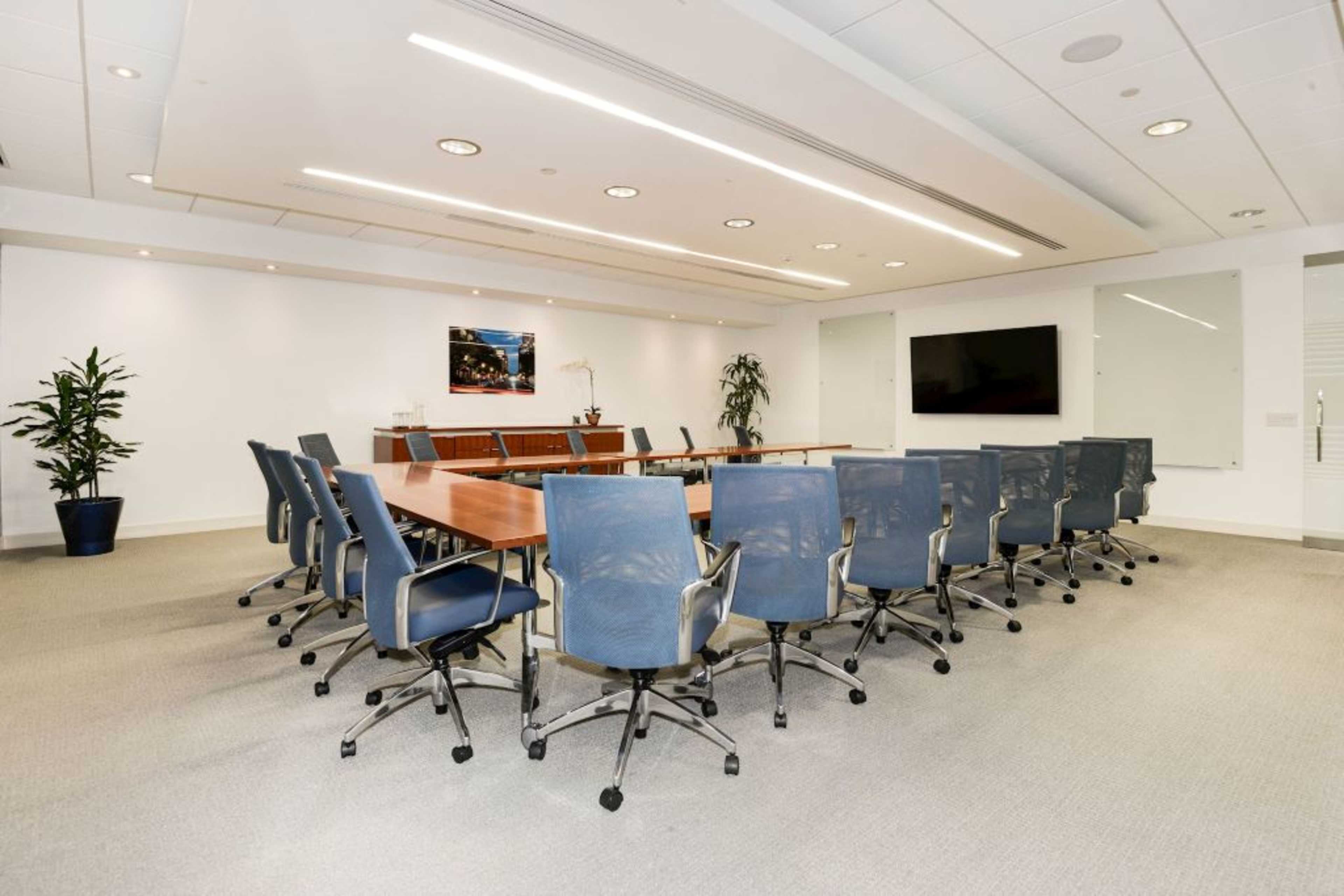 A modern conference room features a long wooden table surrounded by ergonomic chairs, with a large screen and potted plants in the background.