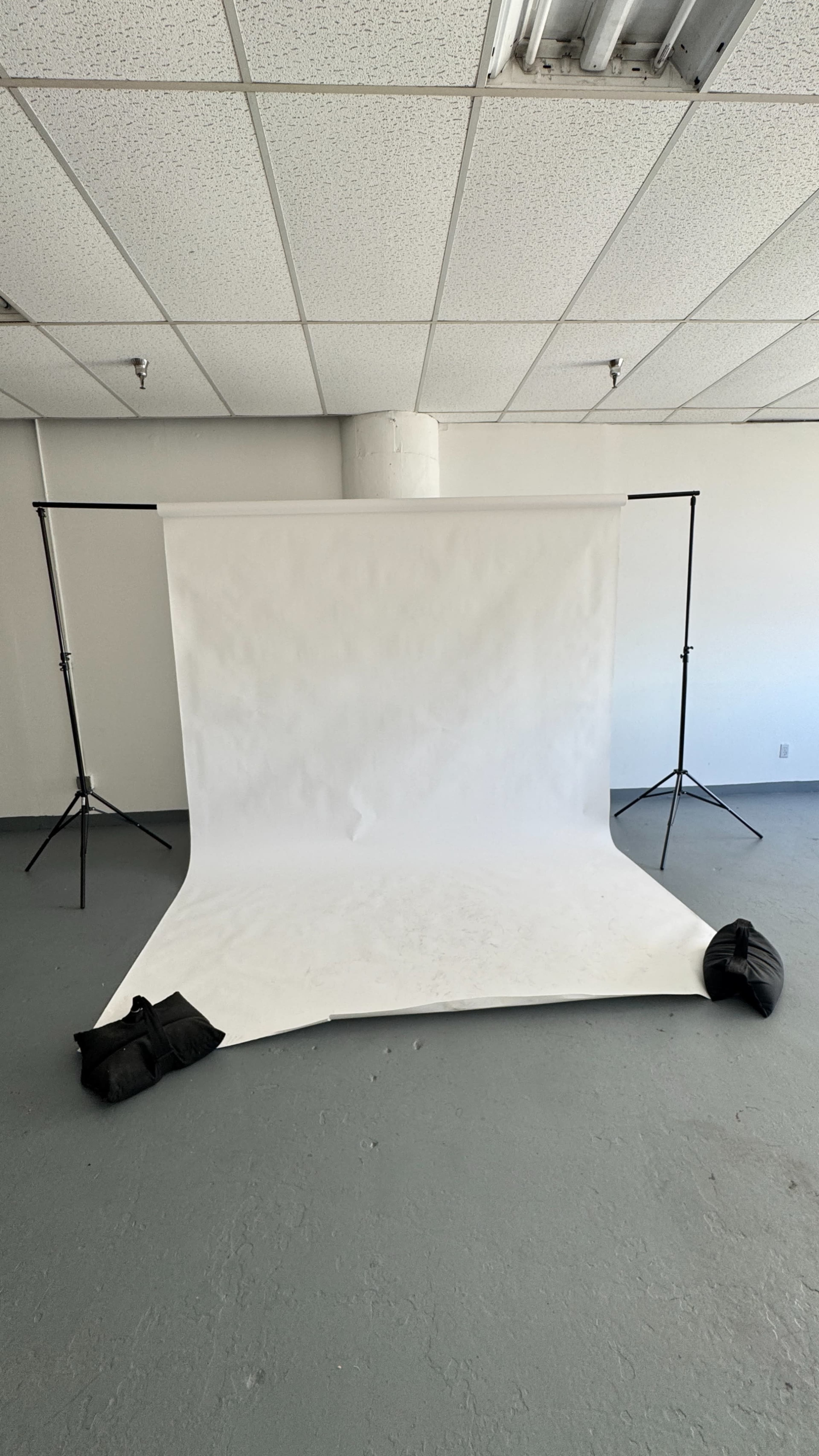 The image shows a photography studio with a white backdrop set up on a gray floor, supported by black stands and weighted down with black bags.