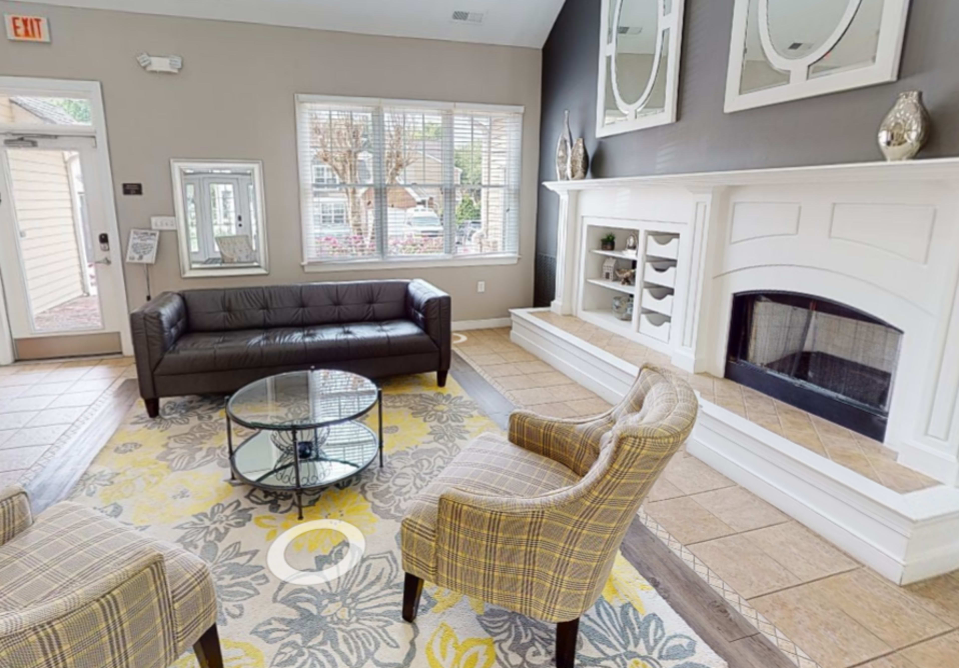 A lobby area featuring a black leather couch, two patterned chairs, a glass coffee table, and a fireplace with shelves, all set against a wall with mirrors.