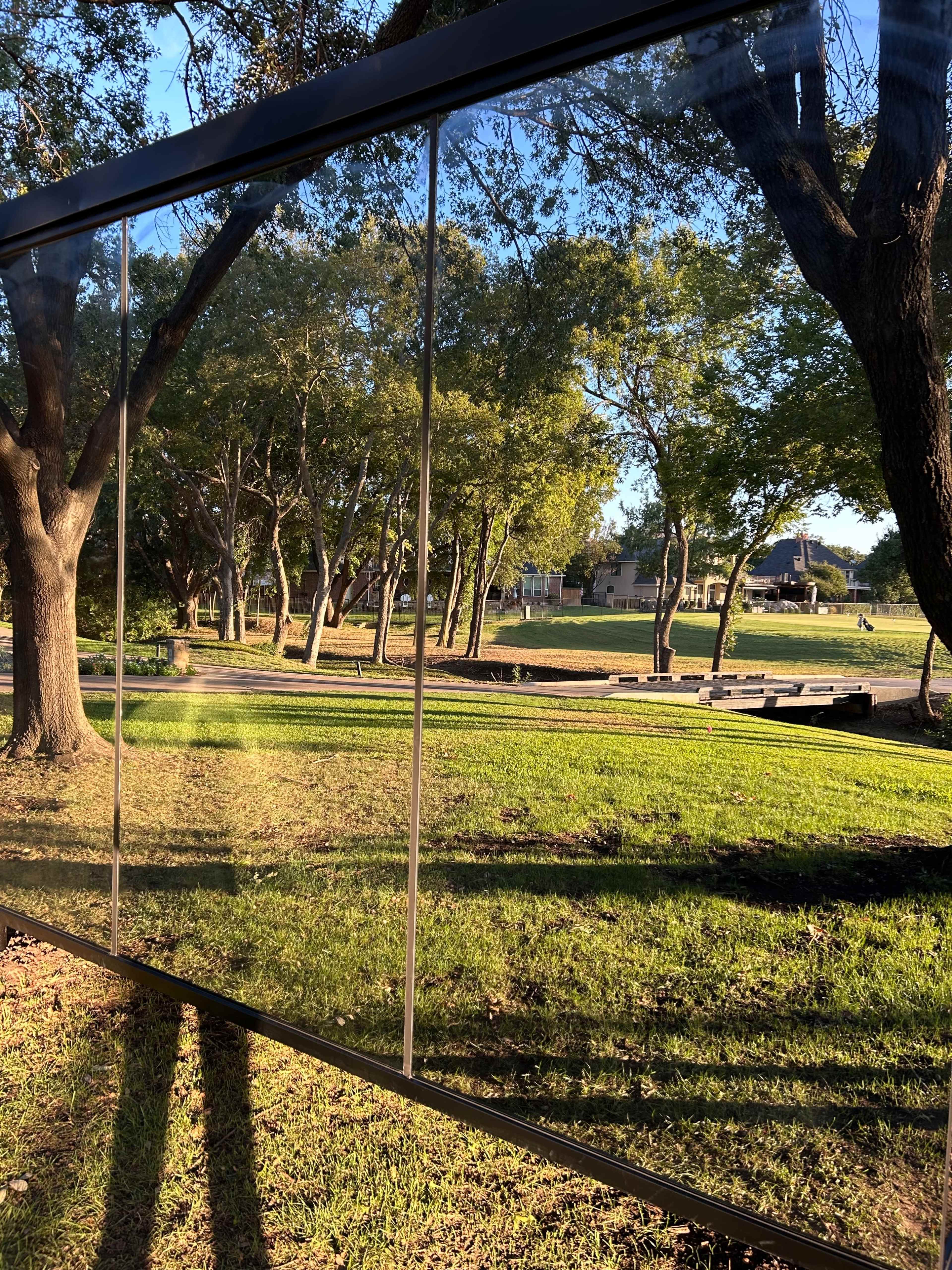 A reflective glass panel stands in a park, mirroring trees and a grassy area while sunlight casts long shadows on the ground.